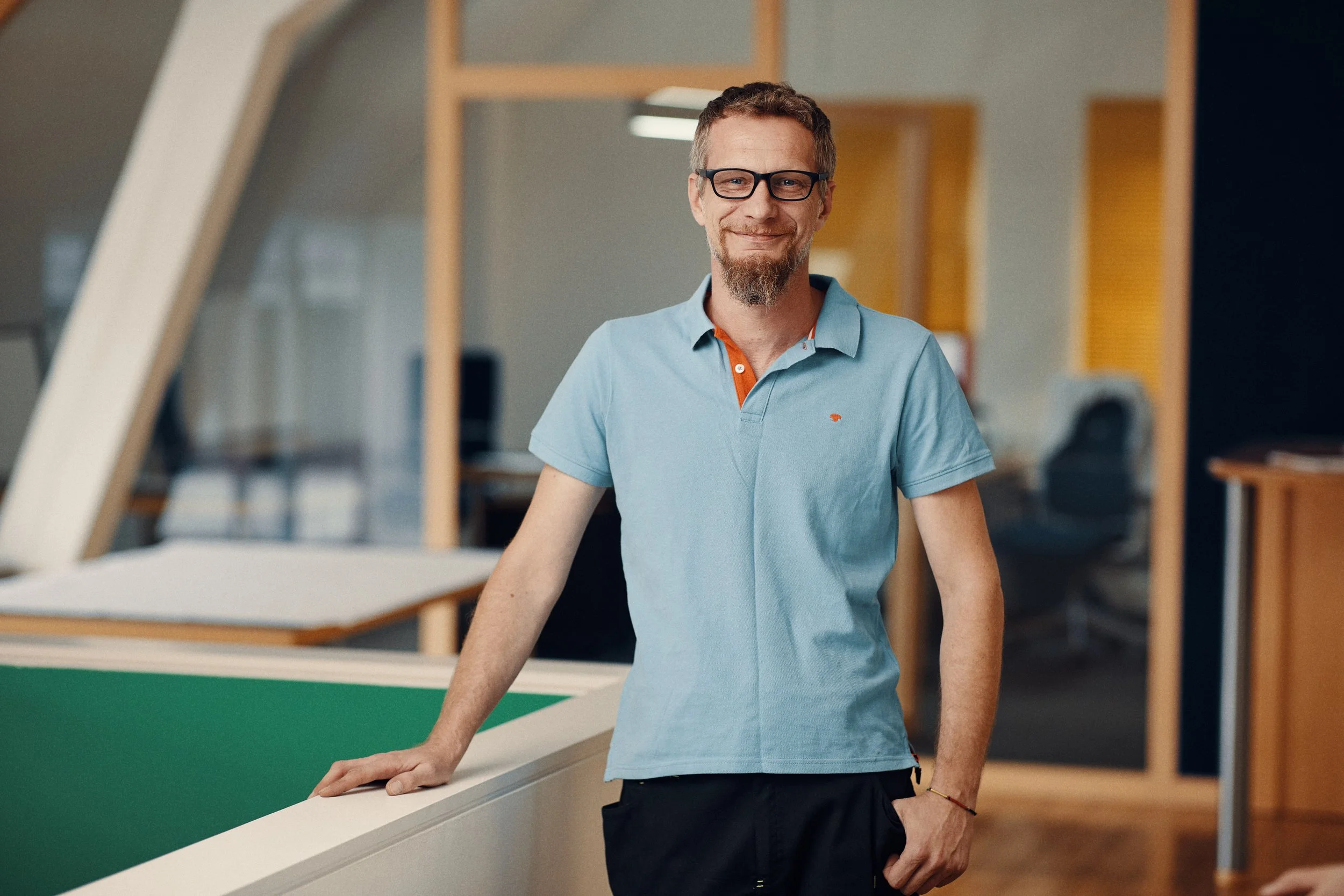Ein lächelnder Mann mit Brille und Bart steht in einem modernen Büroraum, trägt ein hellblaues Polohemd und dunkle Hosen.