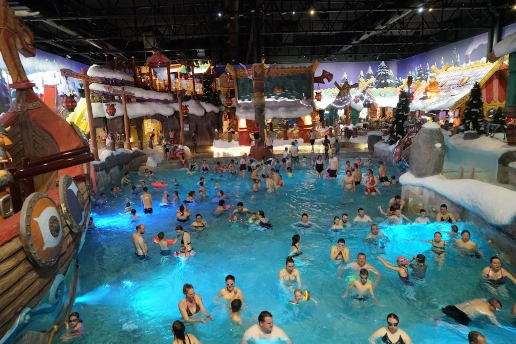 Viele Menschen im Freizeitbad, einige im Wasser, andere am Rand, in einer indoor Wasserlandschaft mit Rutschen und winterlicher Dekoration.