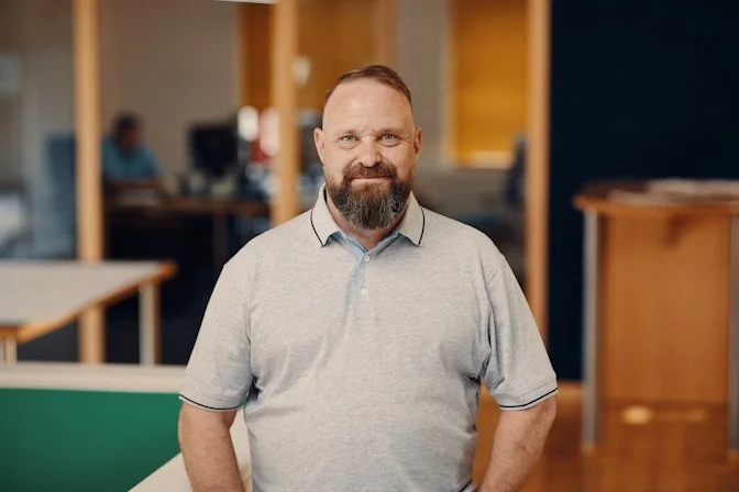 Ein Mann mit Bart und kurzen Haaren trägt ein graues Polohemd und steht in einem modernen Büro. Im Hintergrund sind andere Personen an Computern und Bürotische zu sehen.