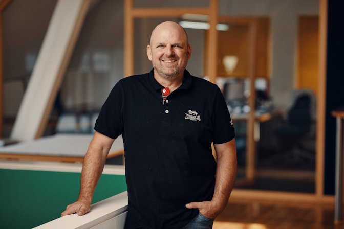 Mann mit kurzen Haaren und Bart, trägt ein schwarzes Polo-Shirt, steht in einem modernen Büro.
