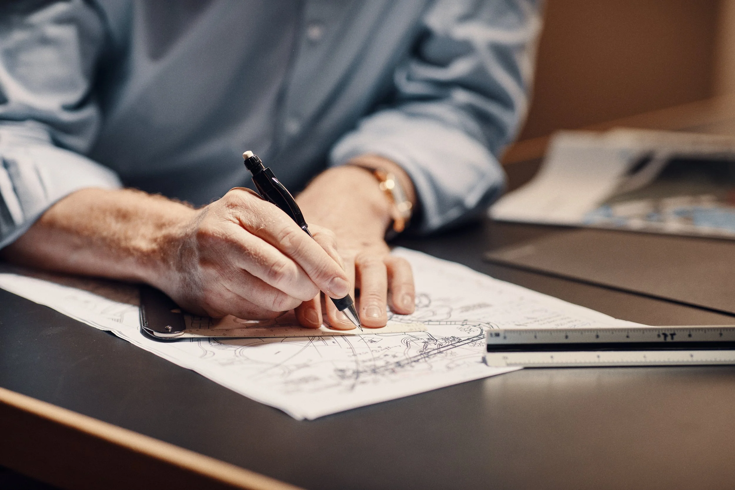 Eine Person zeichnet technische Skizzen auf einem Blatt Papier mit einem schwarzen Stift. Der Tisch enthält ein Lineal, ein Smartphone und einige Ausdrucke.