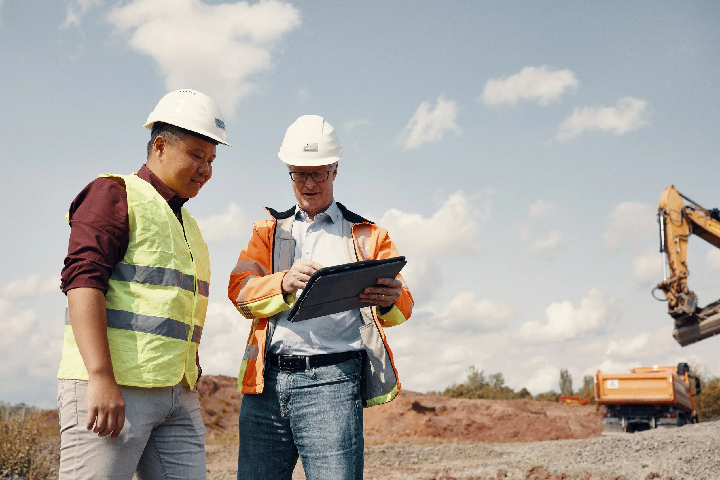 Zwei Bauarbeiter mit Schutzhelmen und Sicherheitswesten stehen auf einer Baustelle und schauen auf eine Tabelle, während im Hintergrund Baumaschinen arbeiten.