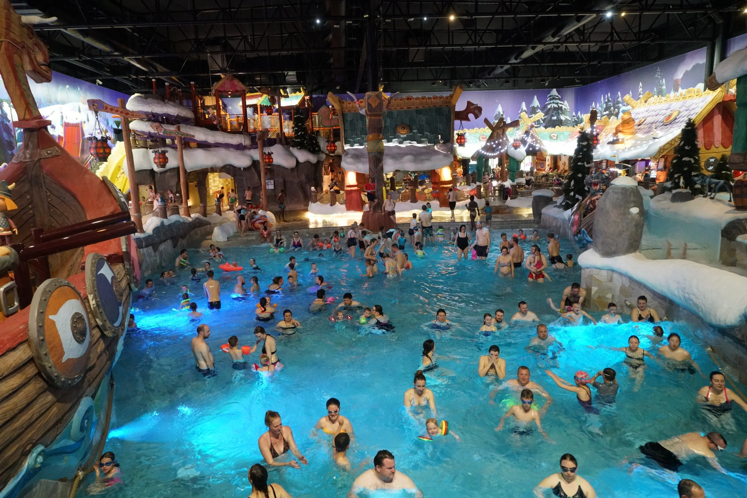 Viele Menschen im Freizeitbad, einige im Wasser, andere am Rand, in einer indoor Wasserlandschaft mit Rutschen und winterlicher Dekoration.
