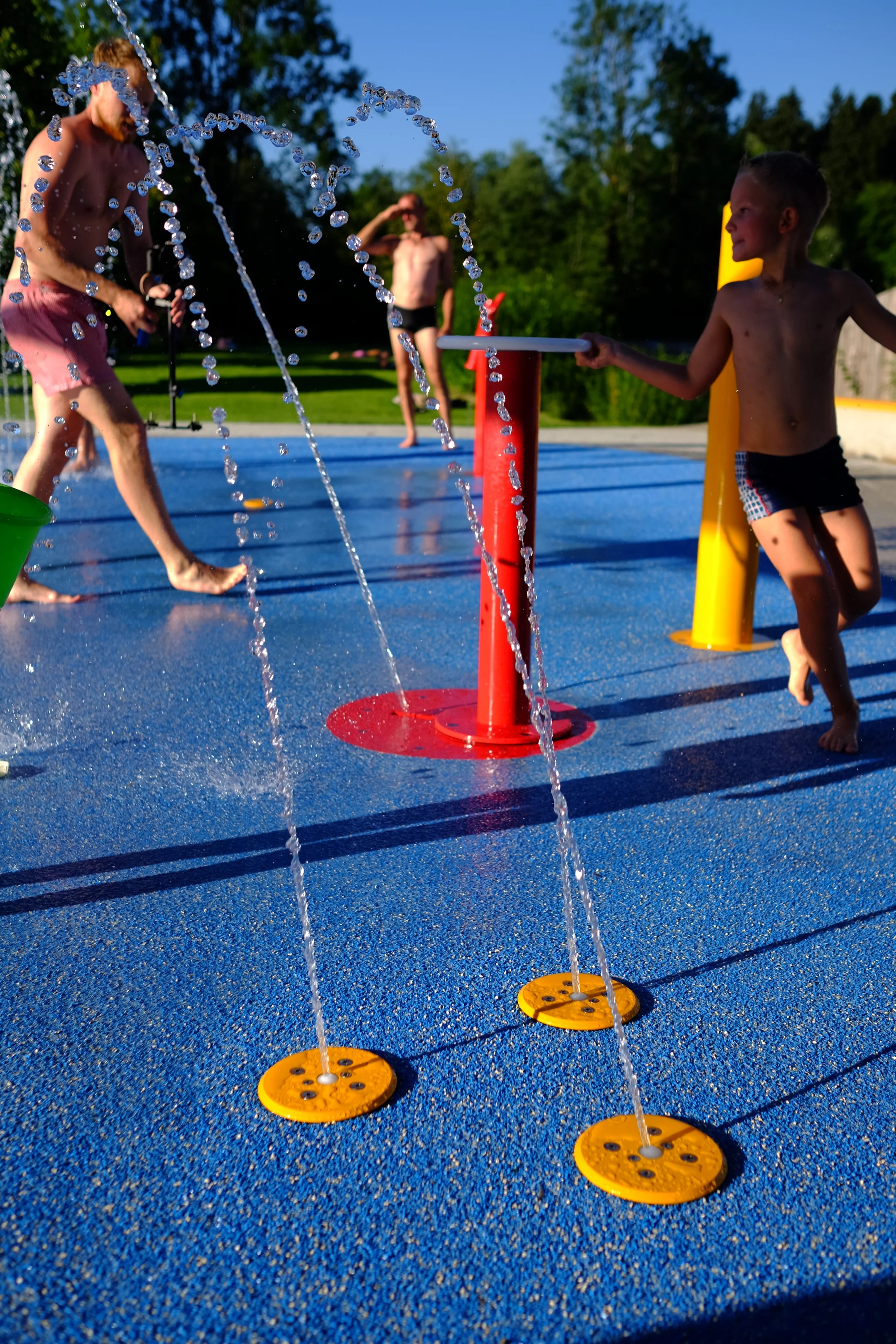 Kinder spielen auf einem Wasserspielplatz bei sonnigem Wetter, während Wasserfontänen auf einer blauen Oberfläche sprühen.