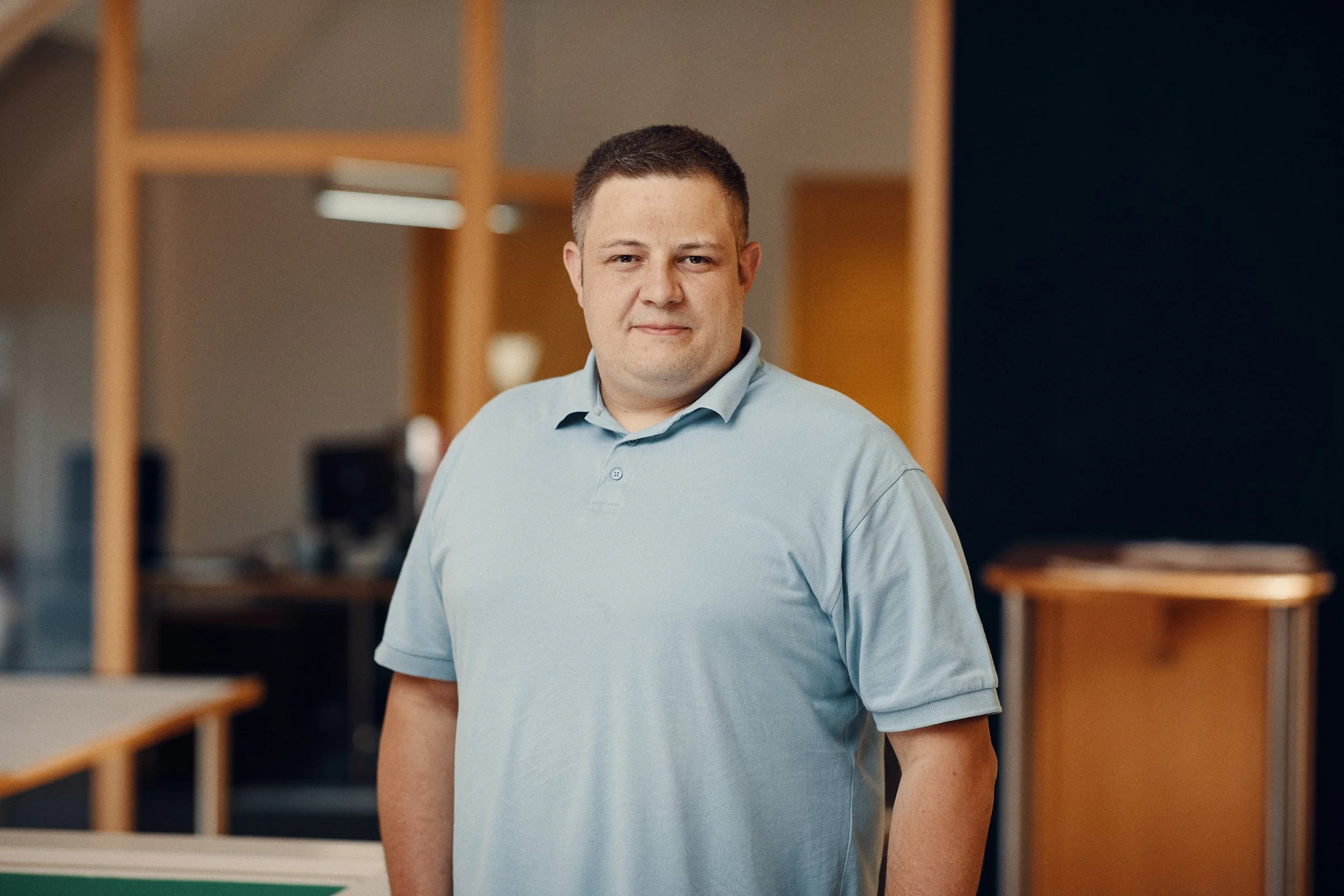 Junger Mann in hellem Polo-Shirt vor einem dunklen Hintergrund, im Hintergrund Holzregale und eine Tabelle.