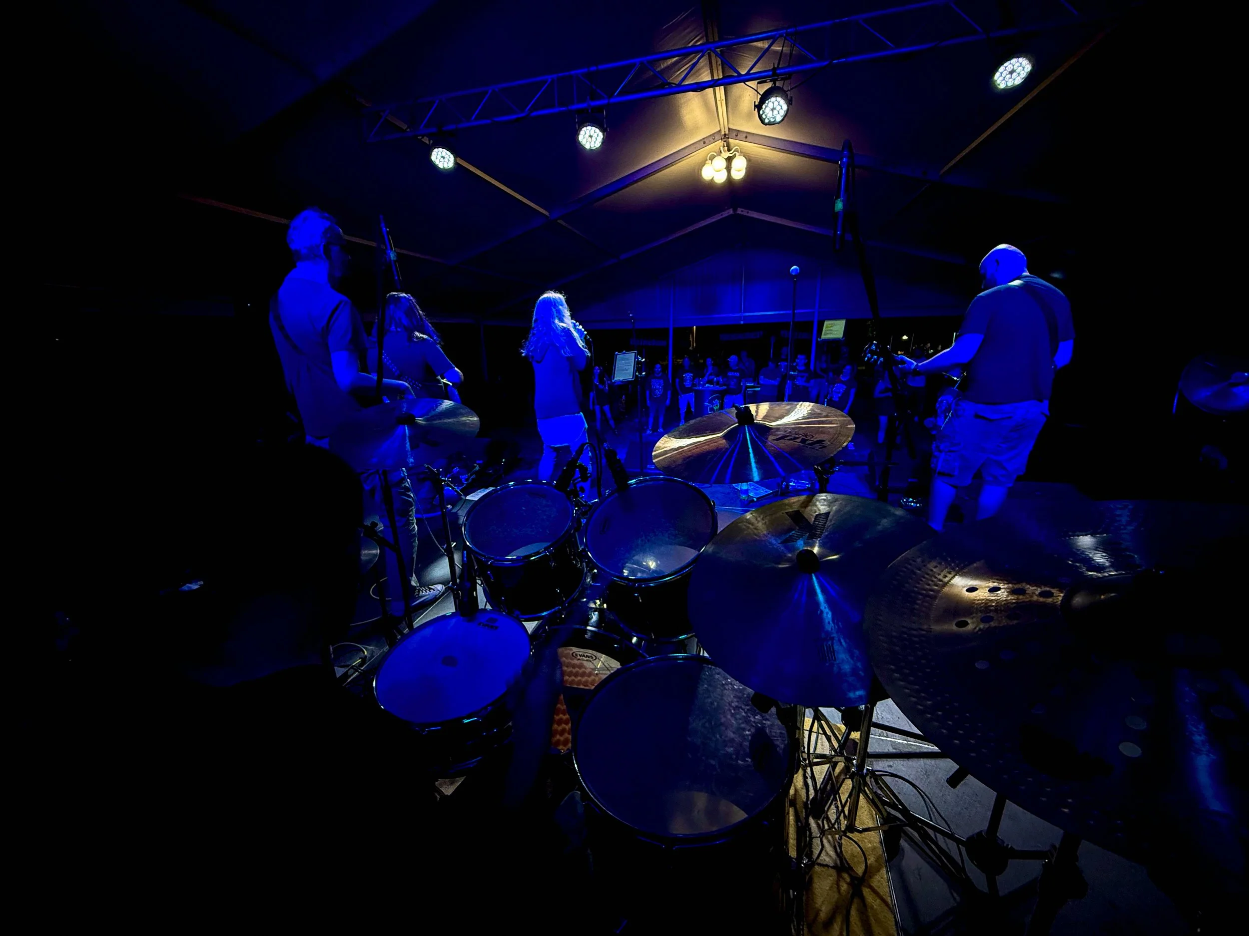 A live band performing under a tent at night, viewed from behind the drum set, with audience in the background.