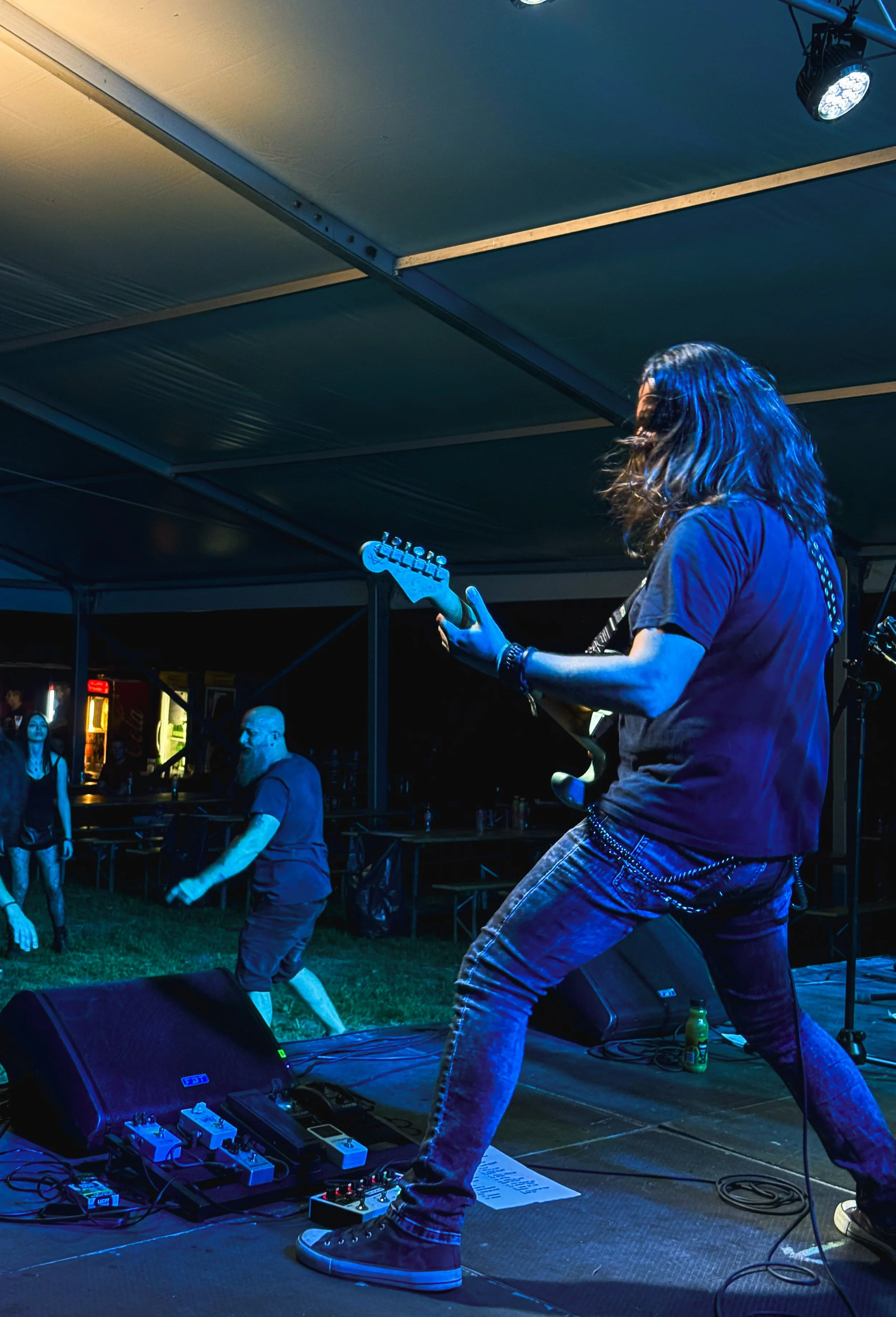 A musician playing electric guitar on stage during a concert at night, with a few audience members visible in the background and stage equipment on the floor.