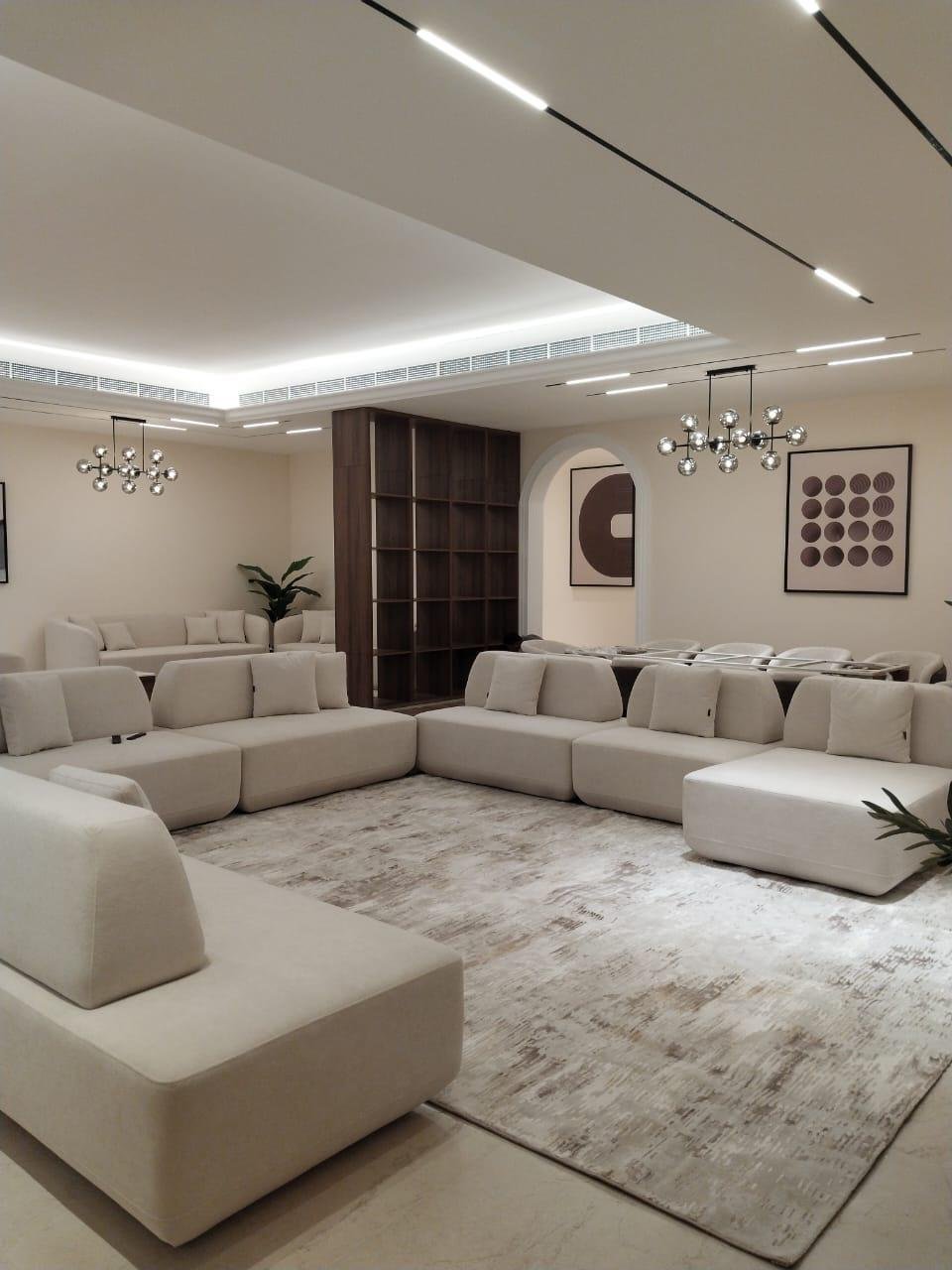Modern living room with beige sectional sofas, abstract artwork, wooden bookshelf, and chrome chandeliers.