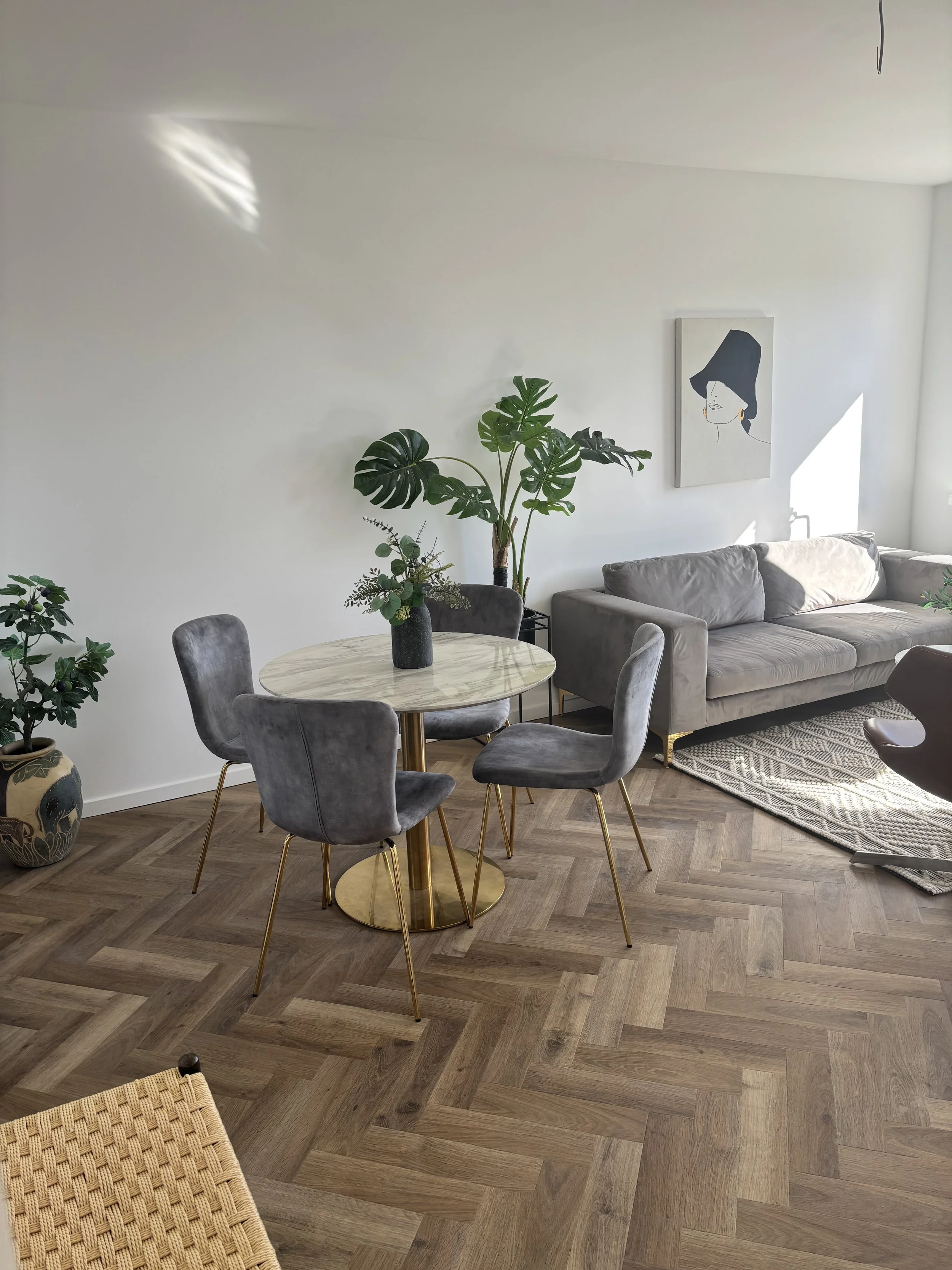 Et moderne stueområde med et rundt spisebord med fire grå stole, en grå sofa, flere grønne planter og en vægkunst af en kvinde med sort hat.