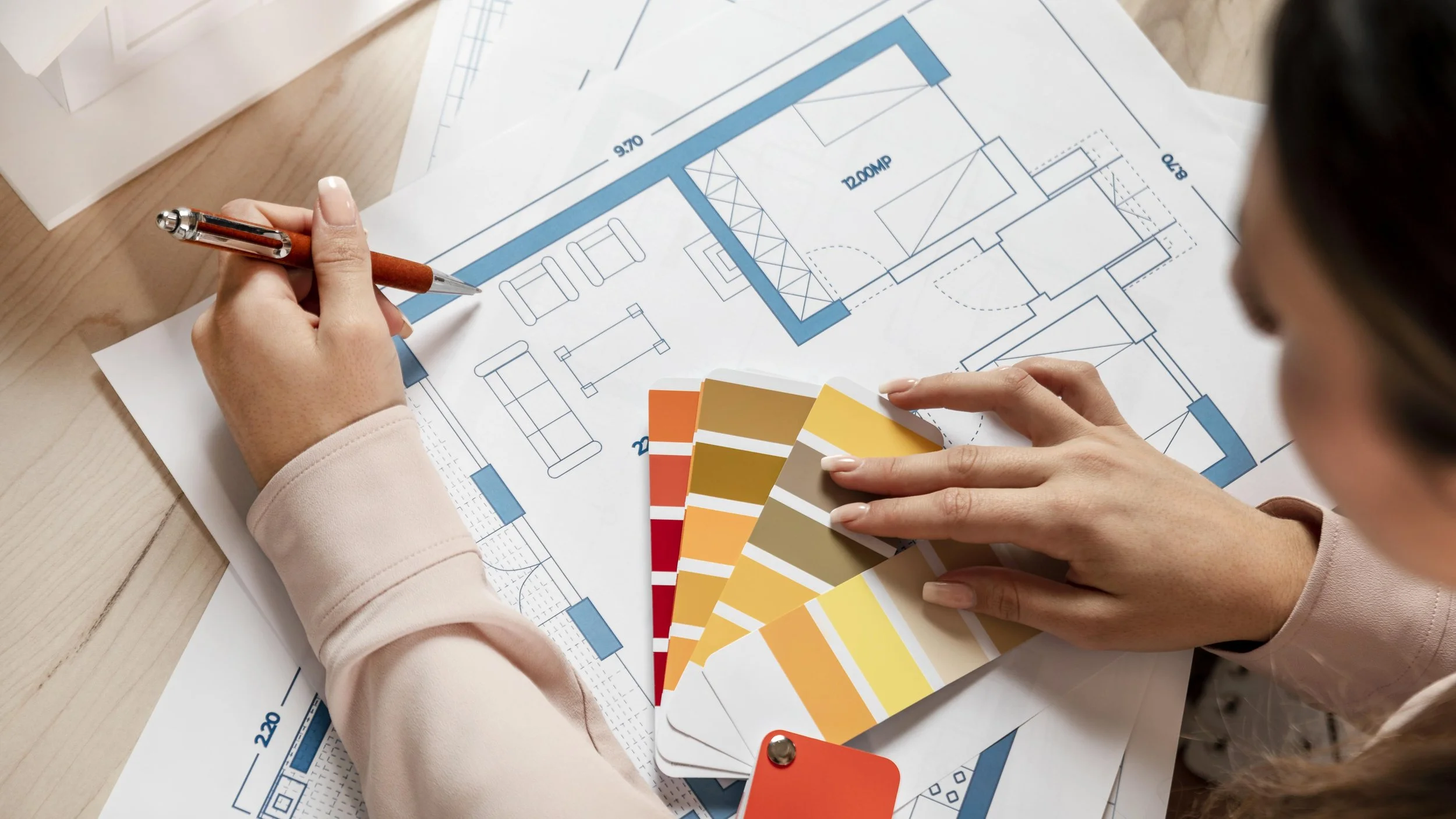 Person planning a house layout with blueprints, color swatches, and a pen on a wooden table.