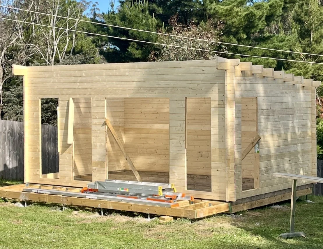 A wooden frame of a small building under construction in a backyard, with construction tools and materials on the ground, surrounded by trees and a wooden fence.
