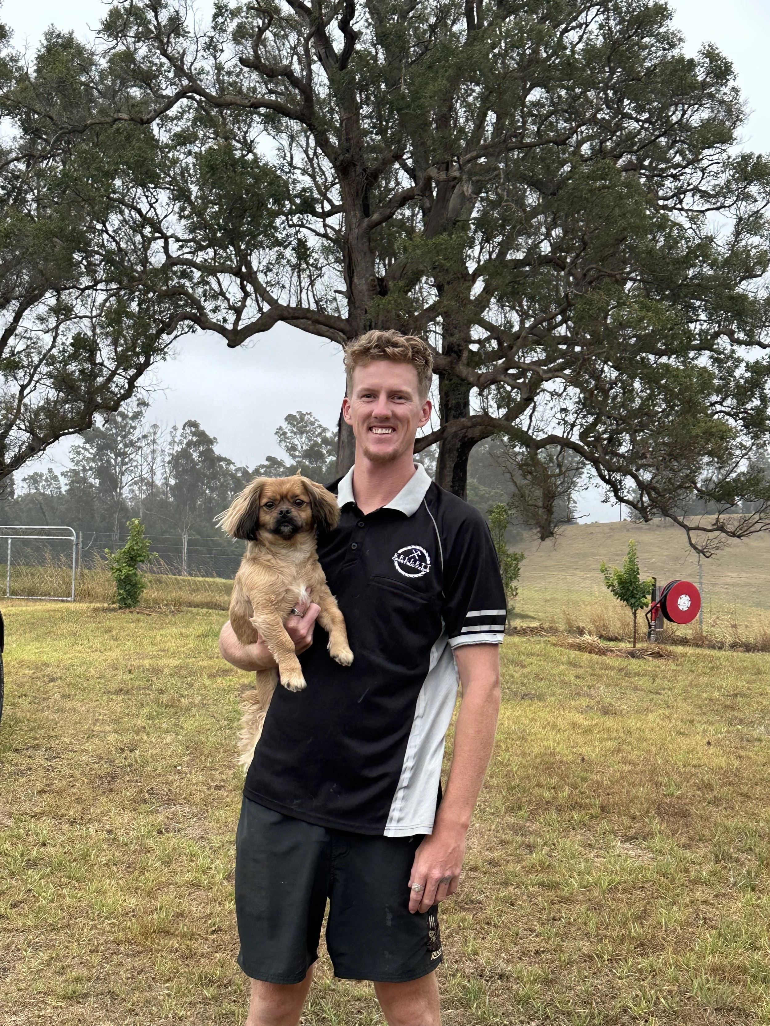 A smiling man holding a small dog in an outdoor grassy area with large trees and a cloudy sky in the background.