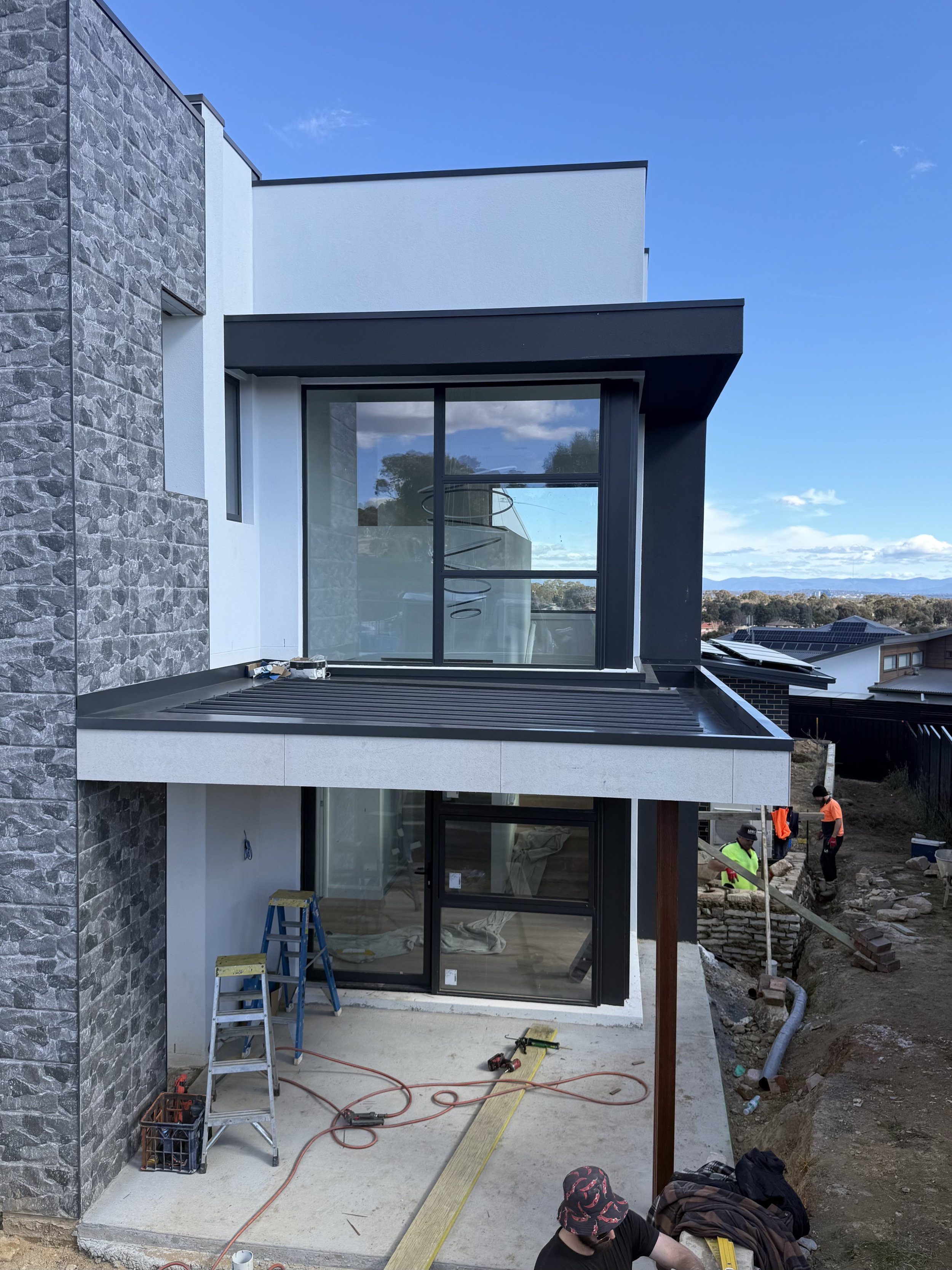 Modern multi-story house under construction, featuring stone and white exterior walls, large glass windows, and a black metal roof. Construction workers are seen working on the site.