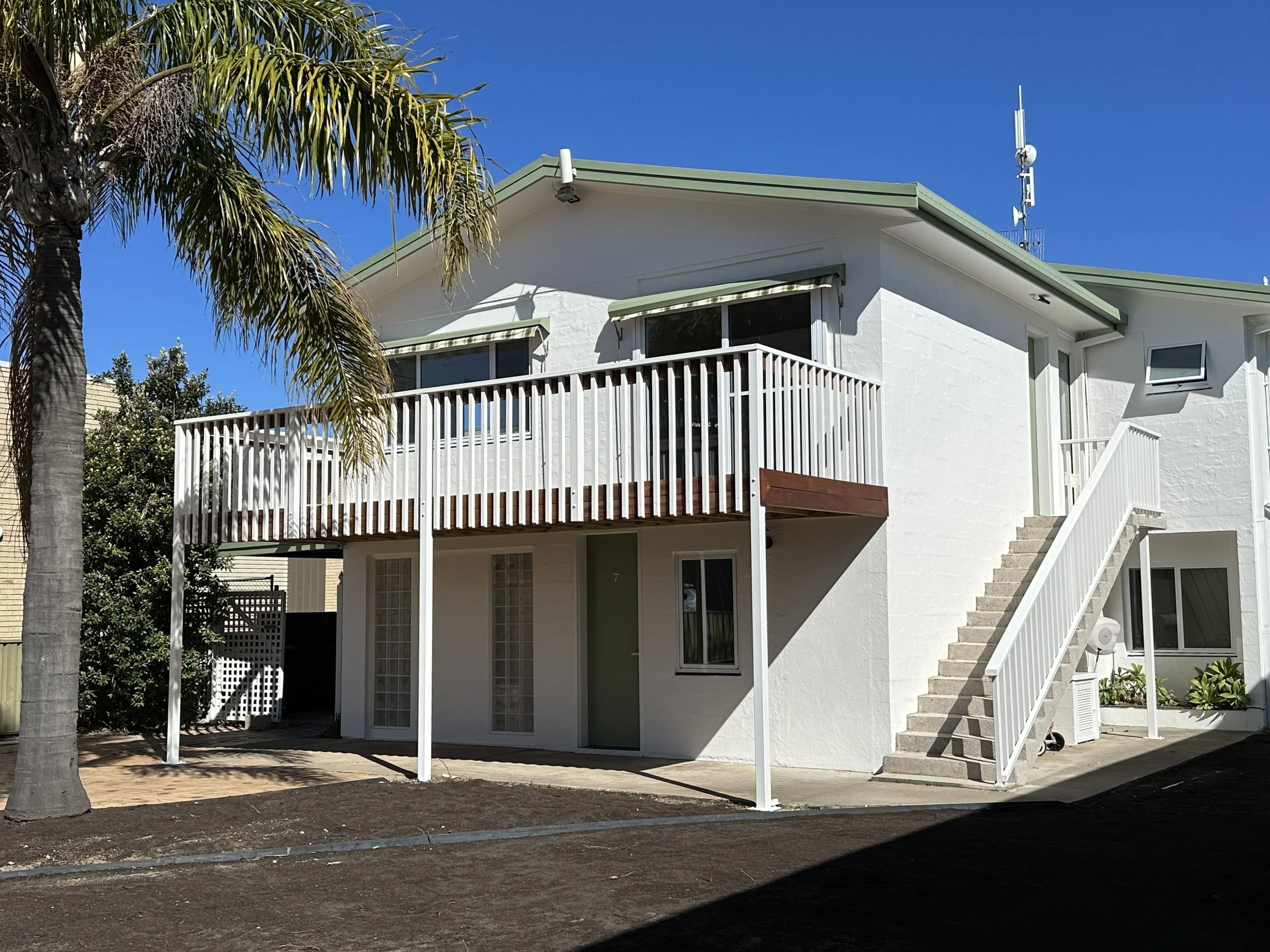 A two-story white house with an exterior staircase, a balcony with a railing, and glass block windows. There are stairs leading to the upper level and a tree with palm leaves on the left.