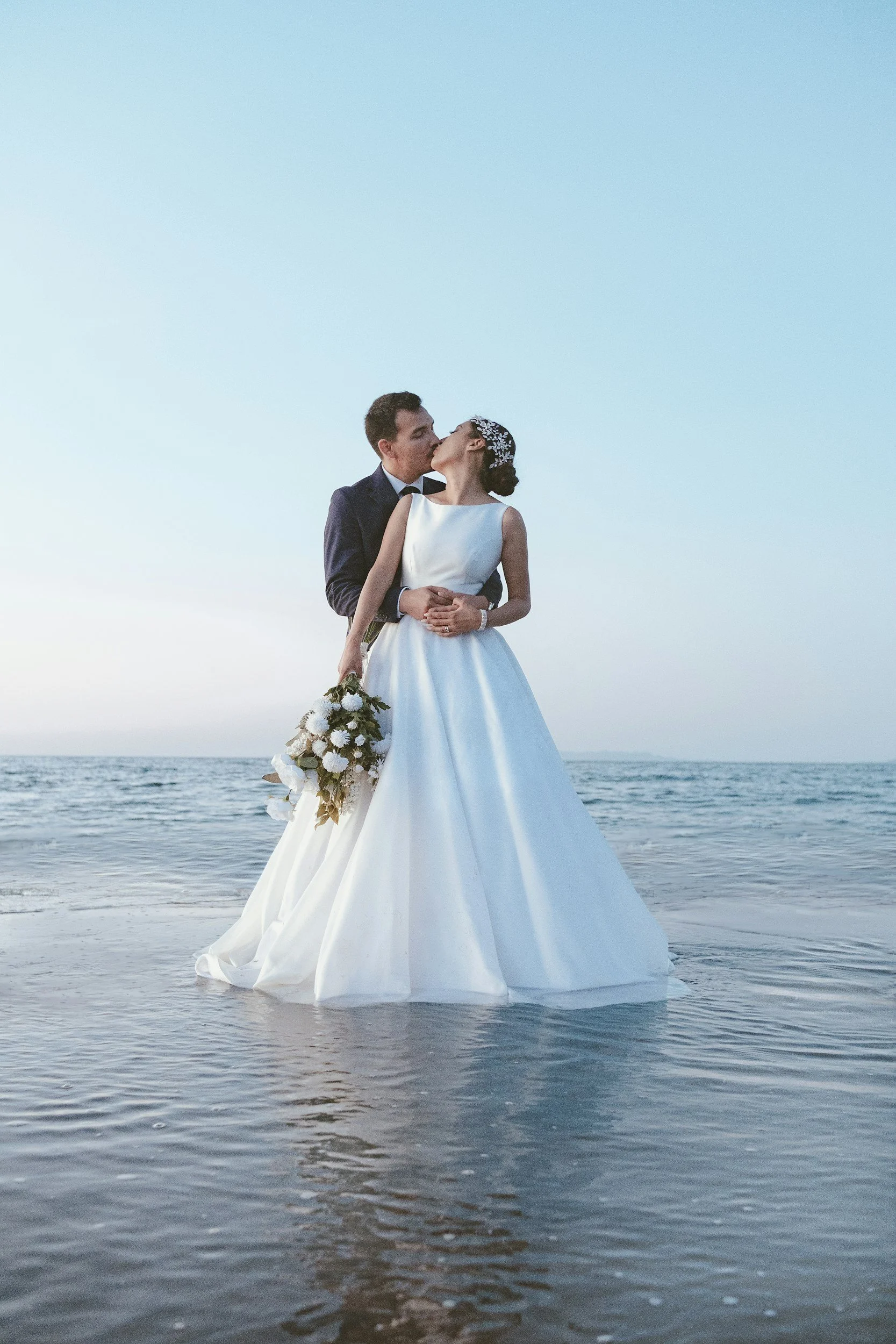 bride and groom embrace each other on the beach with the sun behind them
