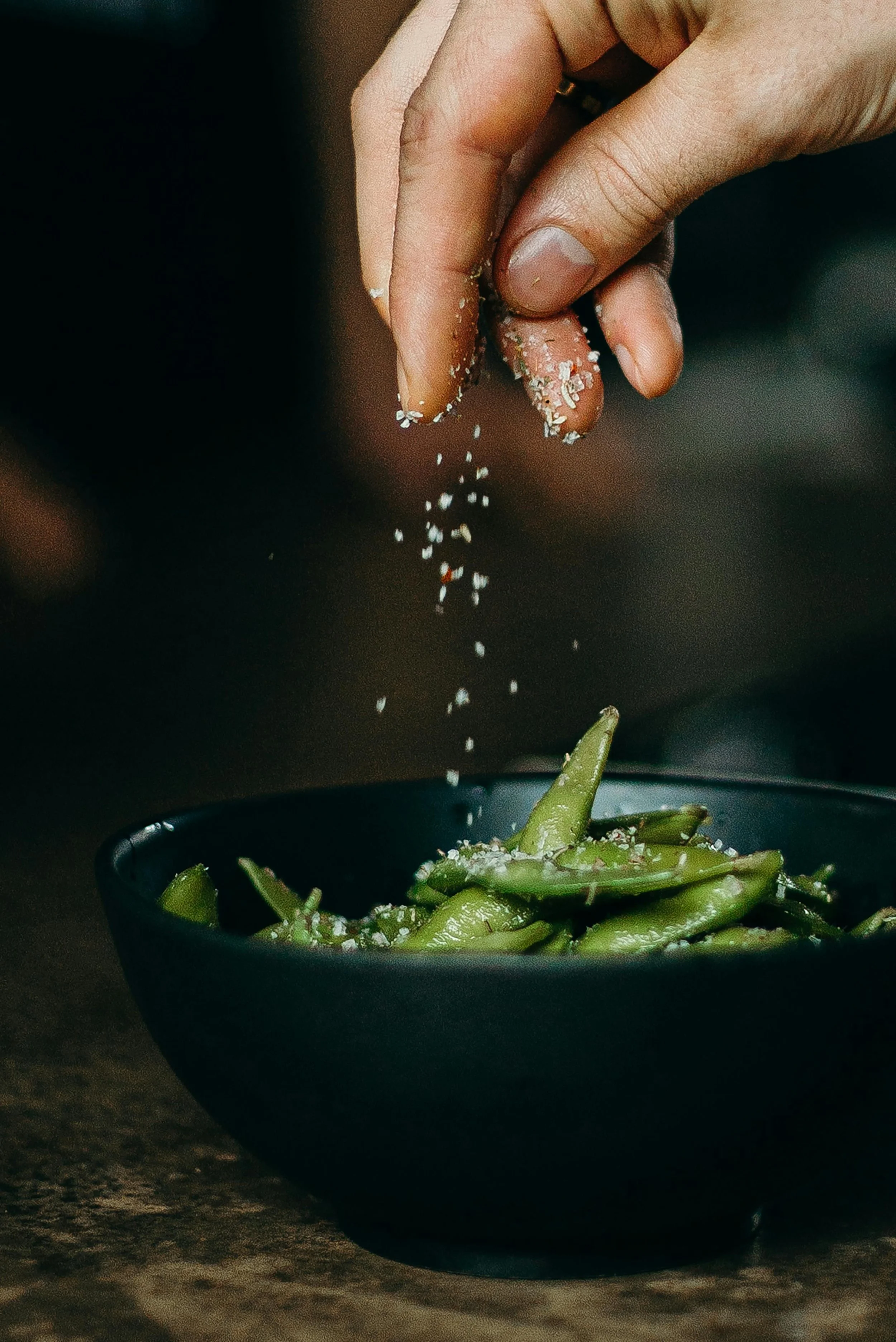 sesame seeds being sprinkled over greens in a black bowl
