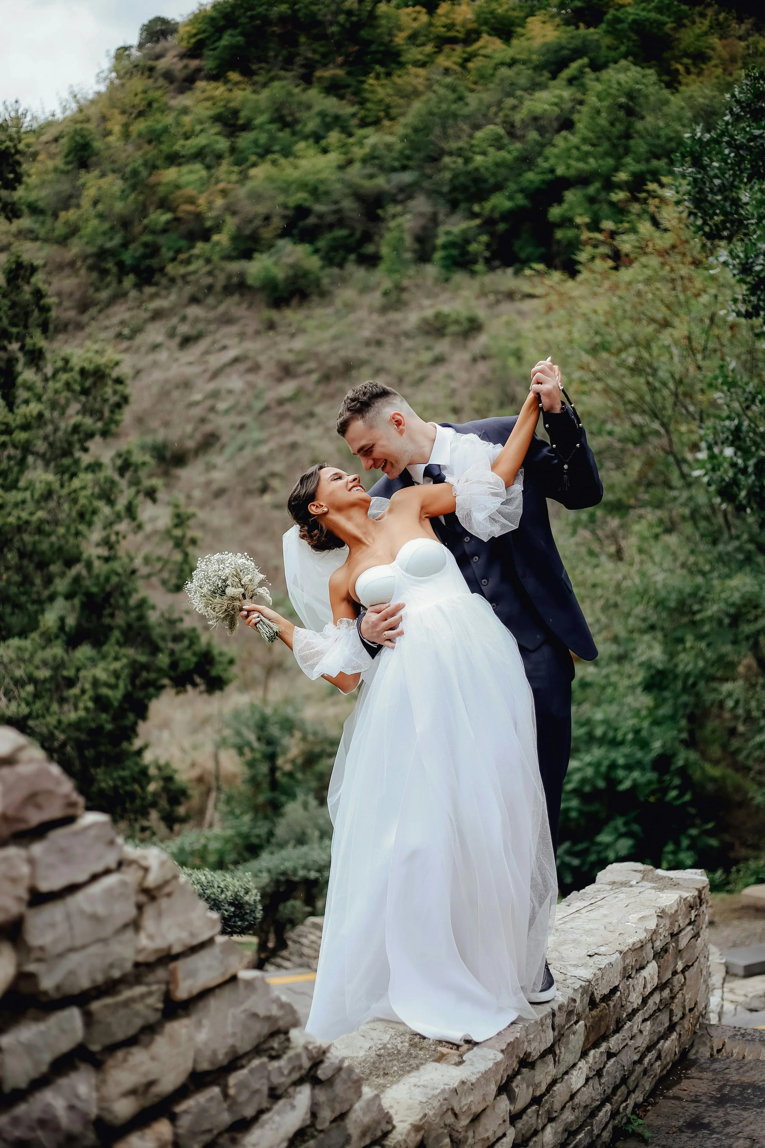 bride and groom stood together on a small narrow wall holding hands and laughing