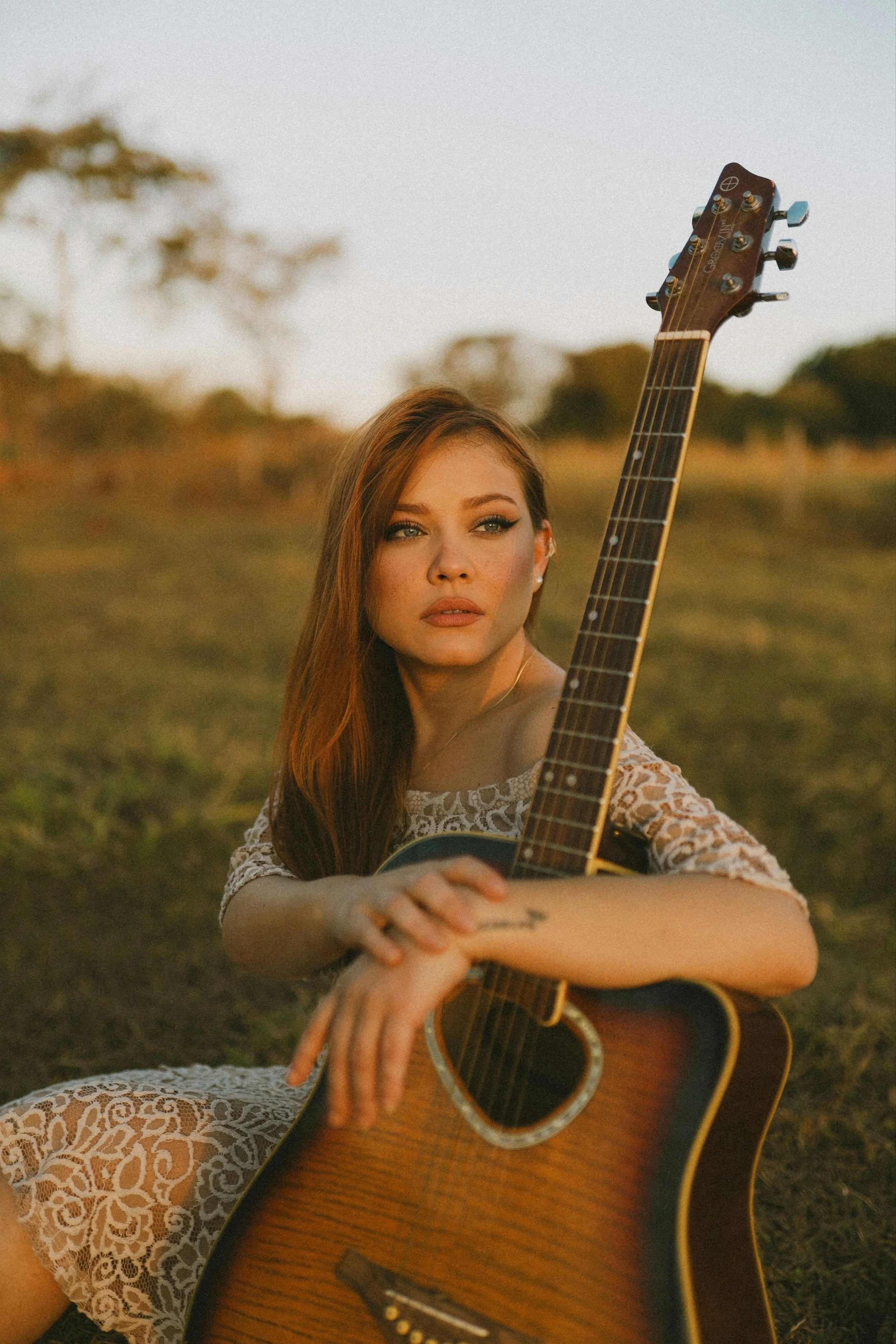 female singer sat in open fields in evening sunlight with guitar
