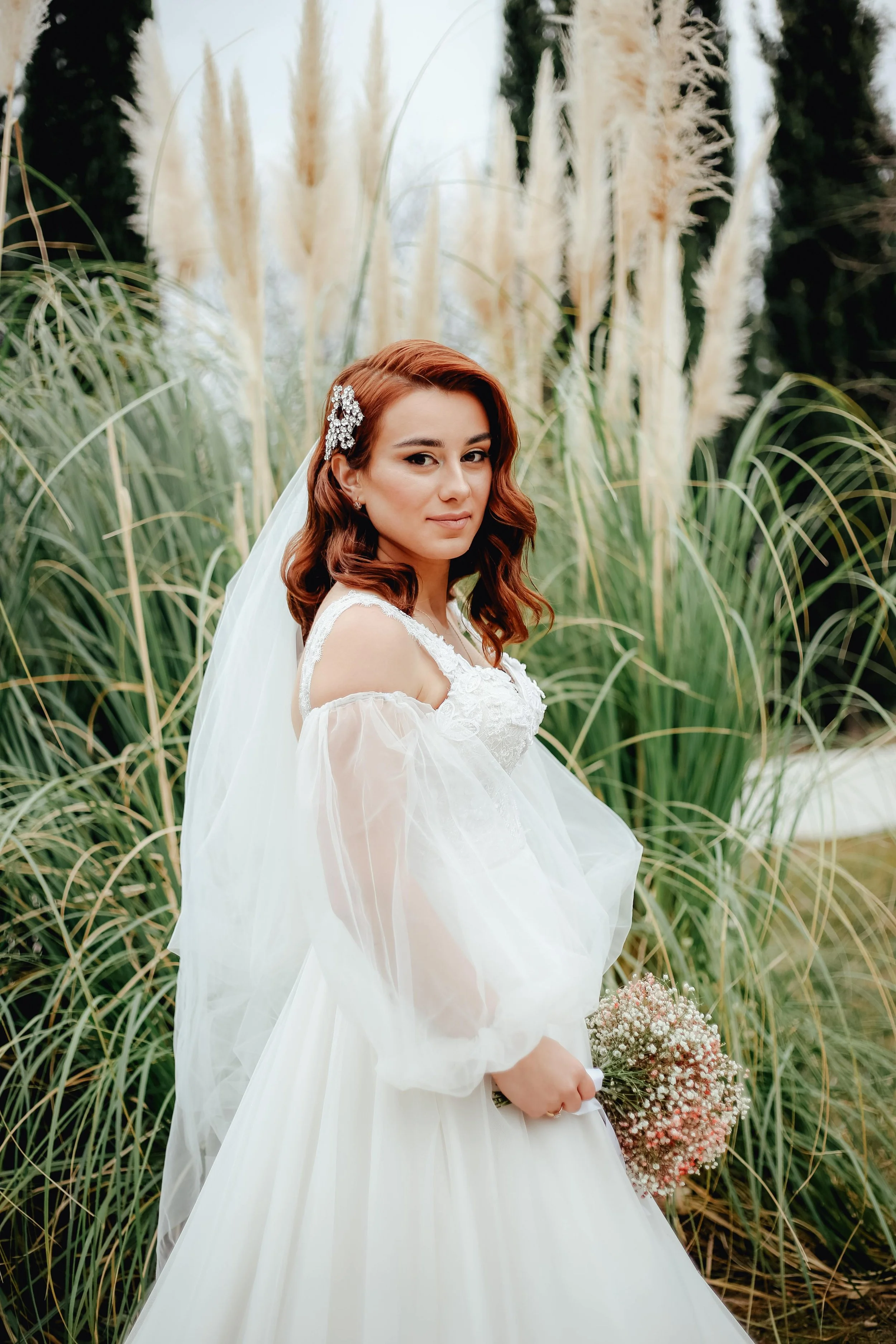 bride with deep red hair holding her wedding flowers, looking at the camera with pampas grass behind her