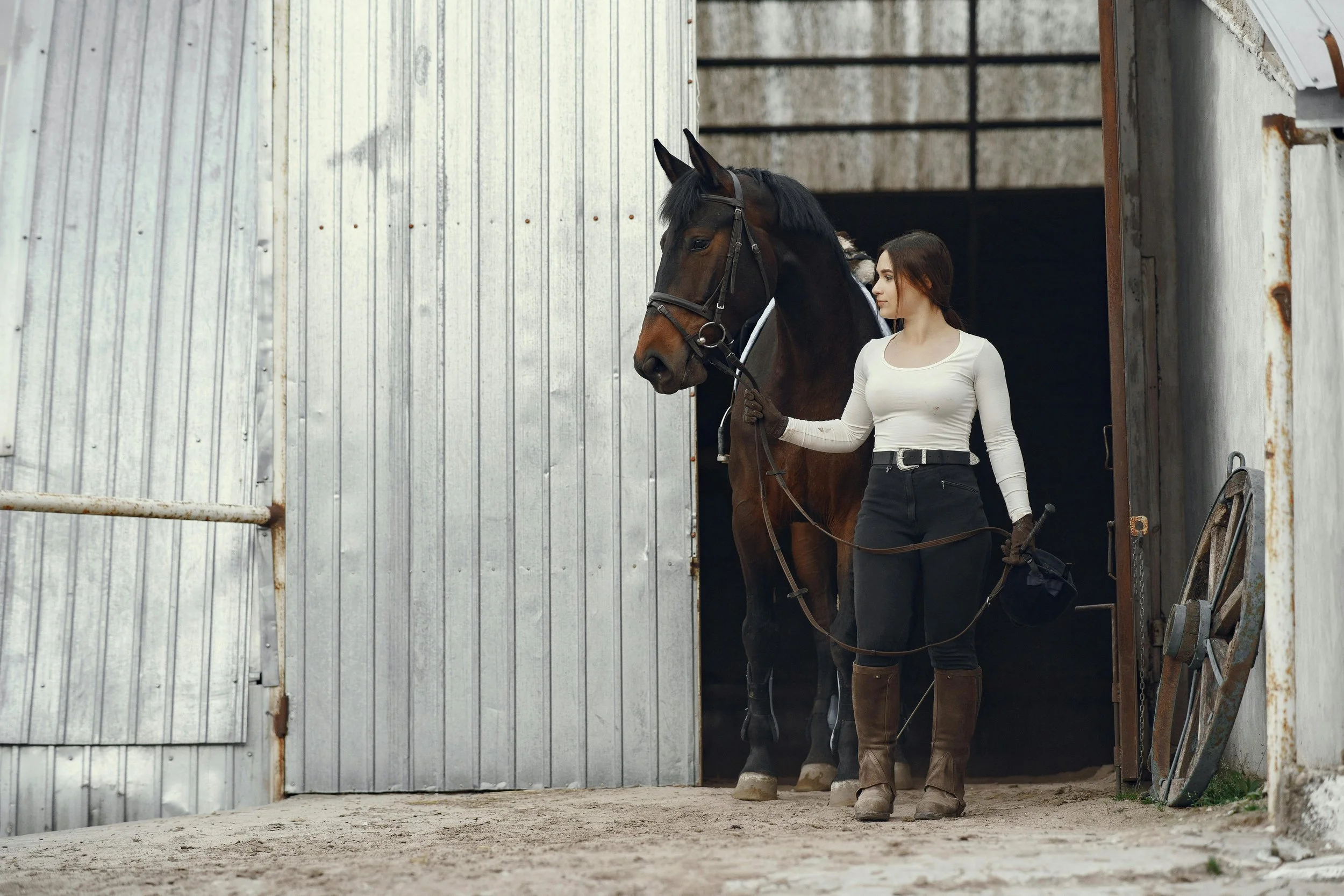 girl with her horse coming out of the barn stood looking to the left with rustic metal behind them