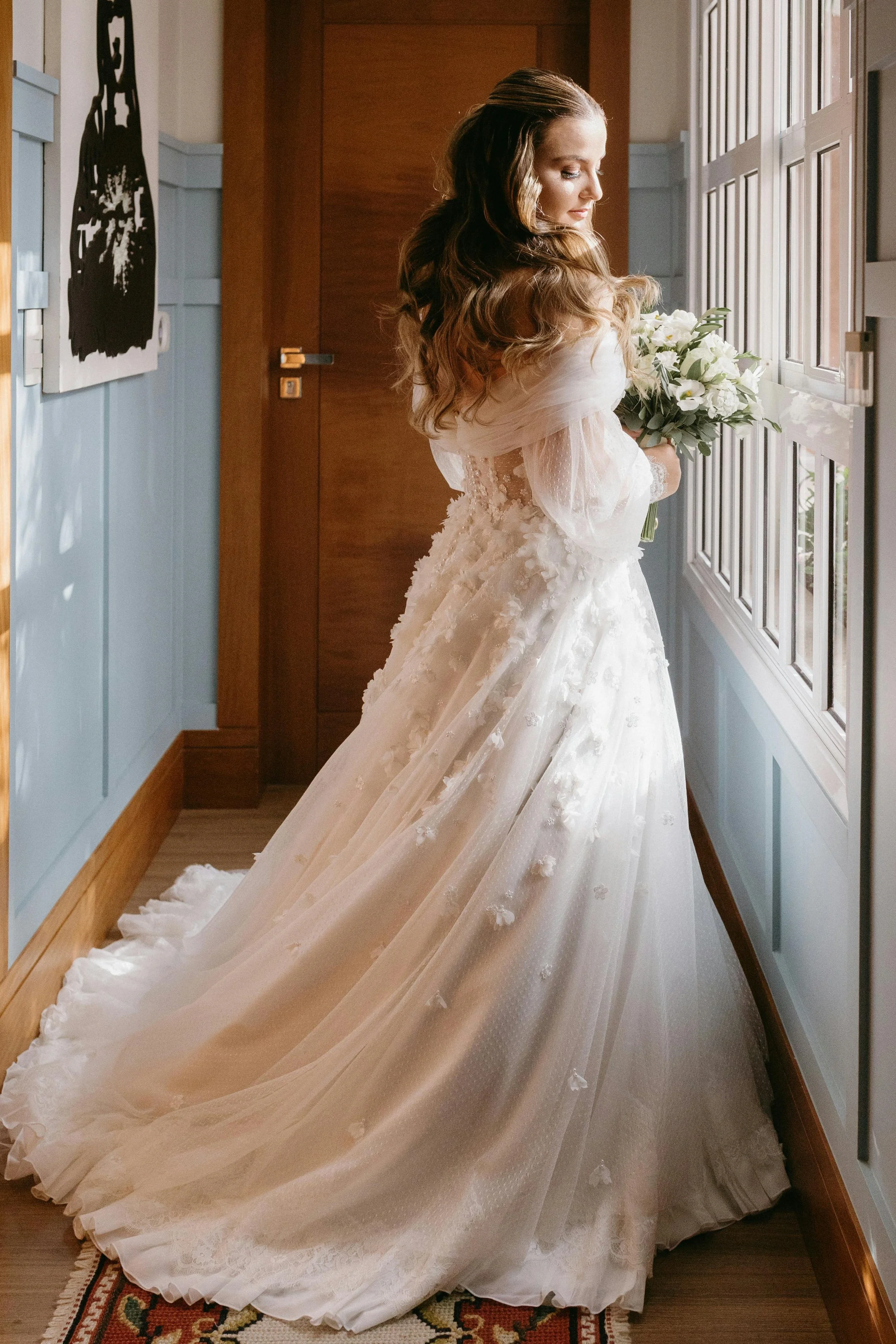 bride ready to go to ceremony stood in hall looking out of the window holding her flowers
