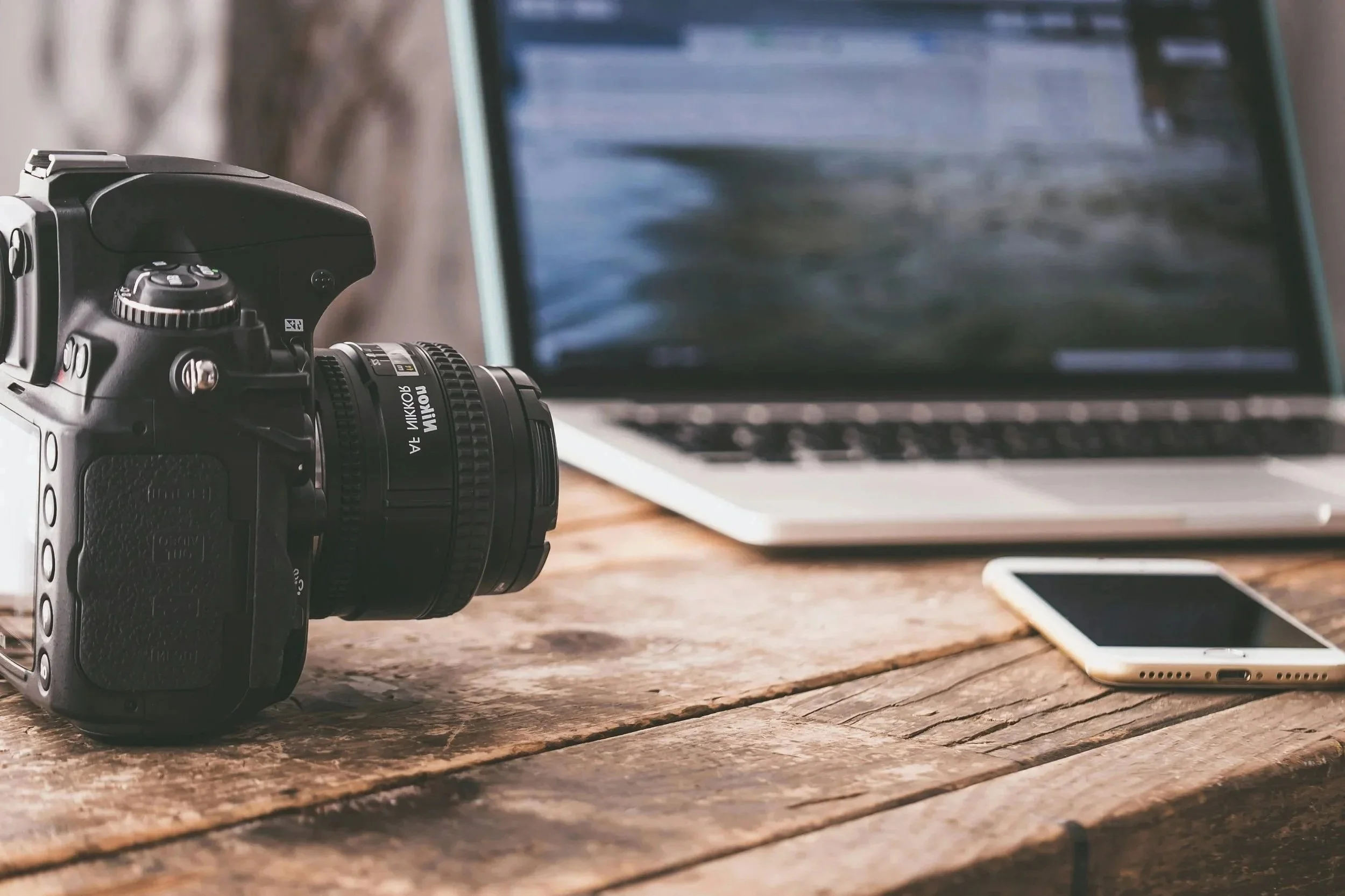 A DSLR camera, a laptop, and a smartphone placed on a rustic wooden table.