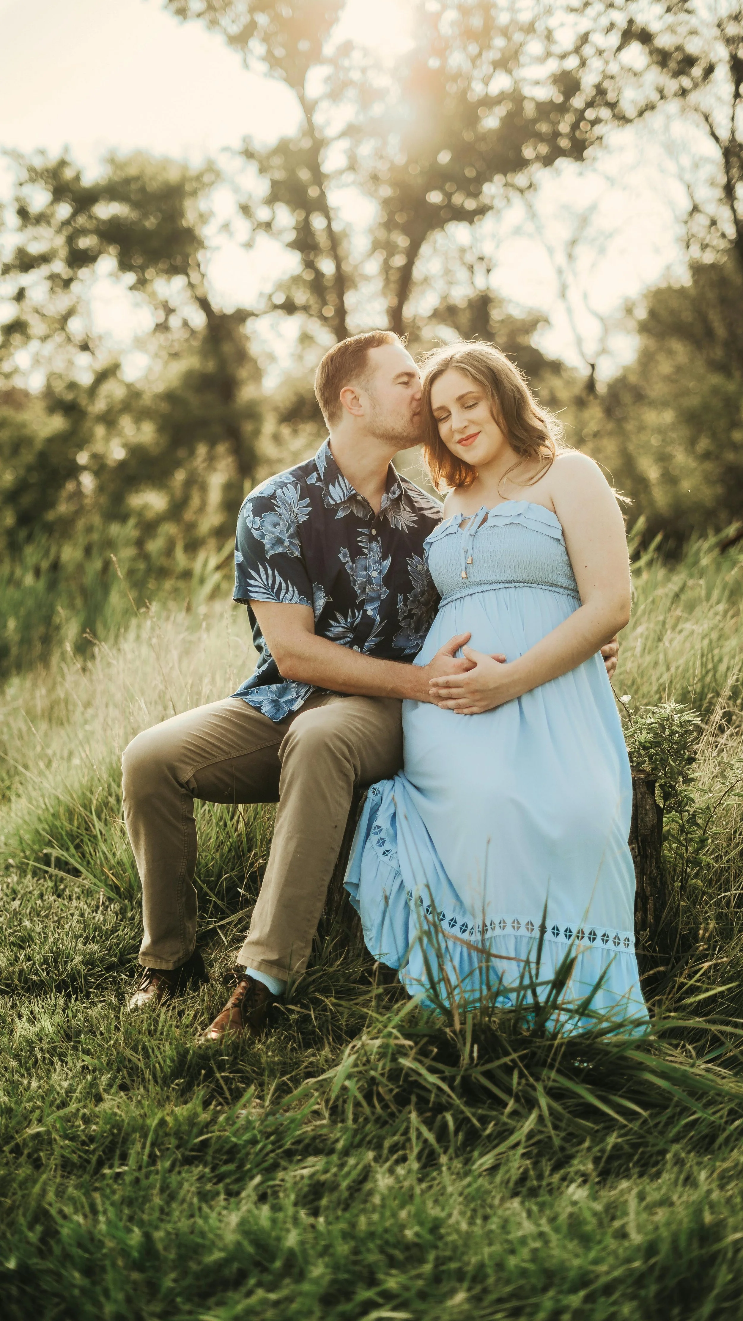 expecting parents sat together in open fields sharing a kiss
