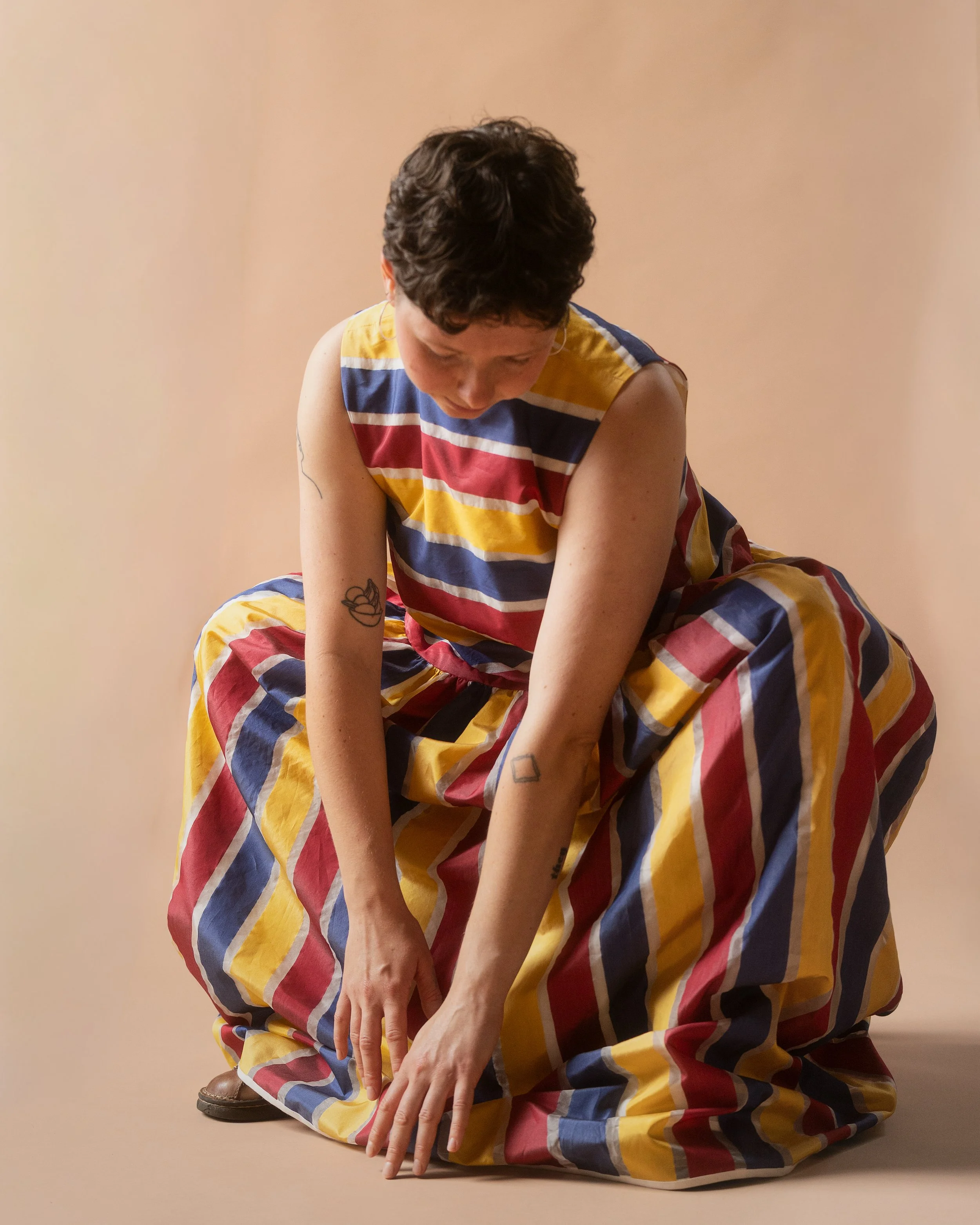 A white person with short brown hair crouches down with their arms outstretched to the floor. They are wearing a blue, yellow, red, and white striped dress made of silk, against a beige background.