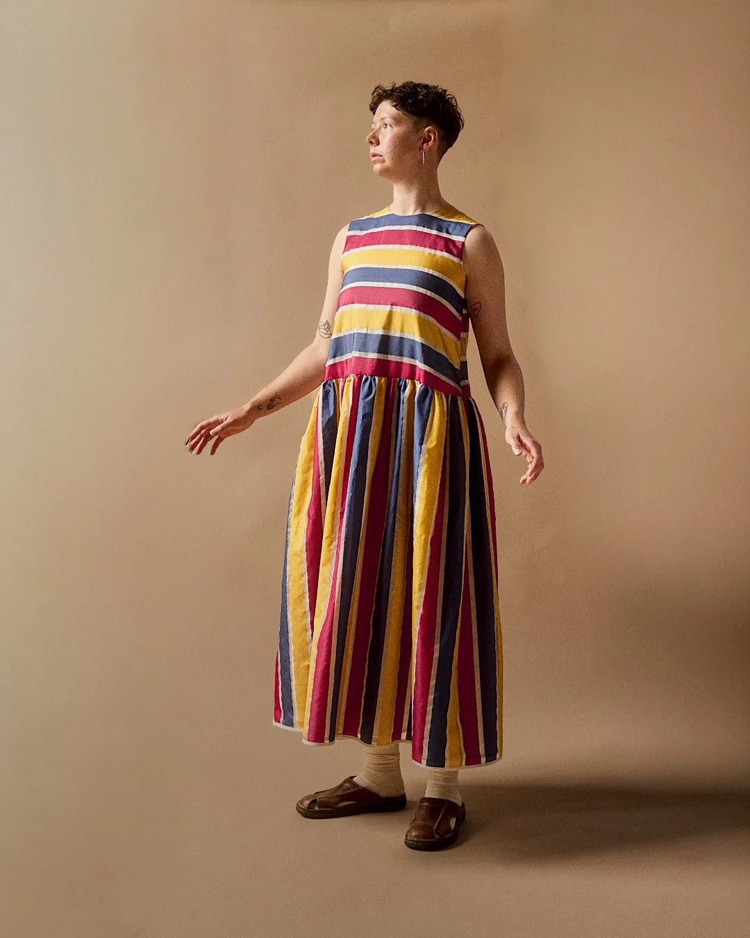 A white person with short brown hair is standing upright with their arms slightly outstretched, looking to their right, against a beige background. They are wearing a blue, yellow, red, and white striped silk dress.