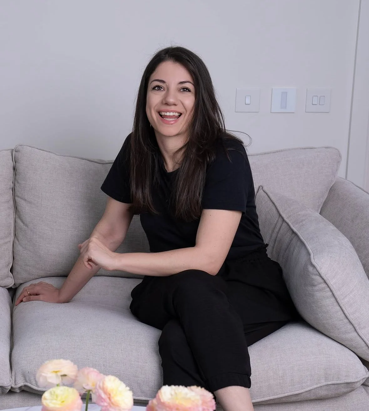 A woman with dark brown hair wearing a black t-shirt and black pants, sitting on a light-colored sofa, smiling and looking at the camera, in a modern living room with light gray walls and visible light switches.