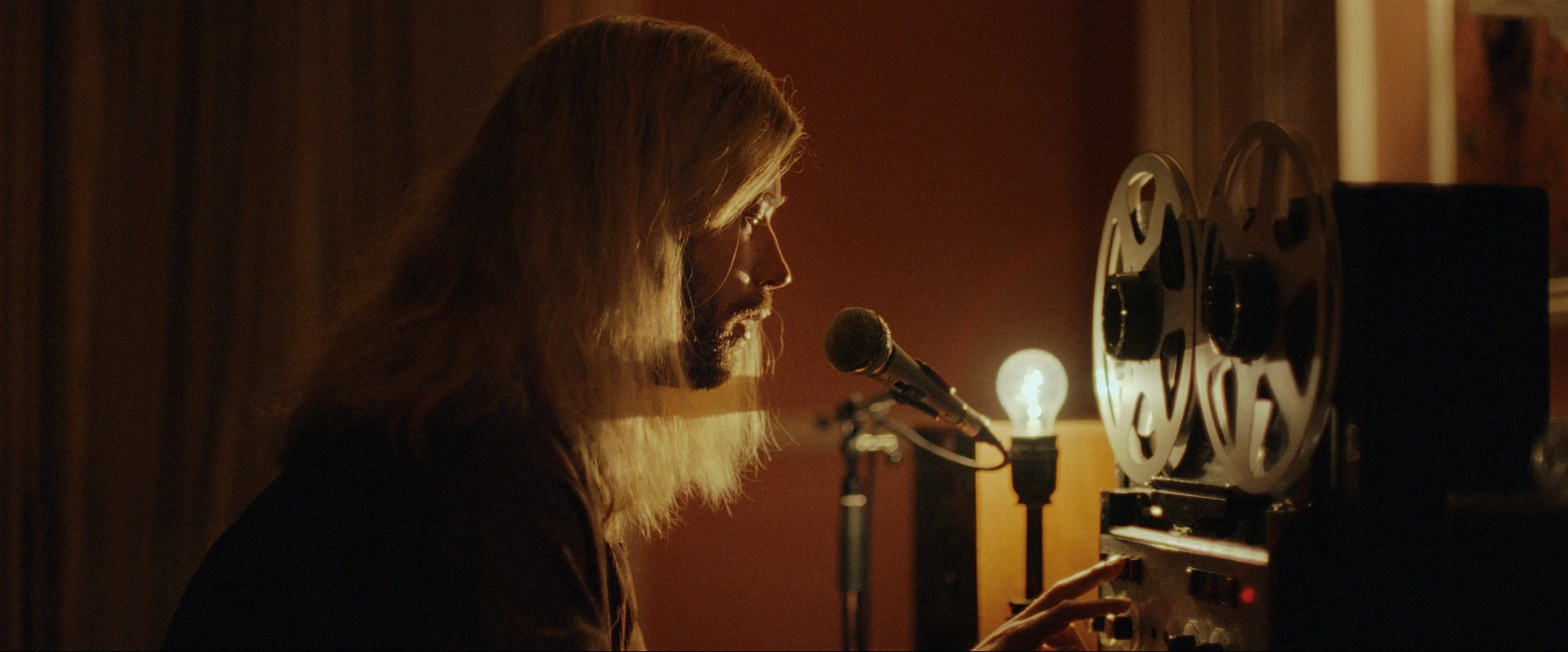 A man with long hair and a beard speaking or singing into a microphone at a recording studio with a reel-to-reel tape recorder and warm ambient lighting.