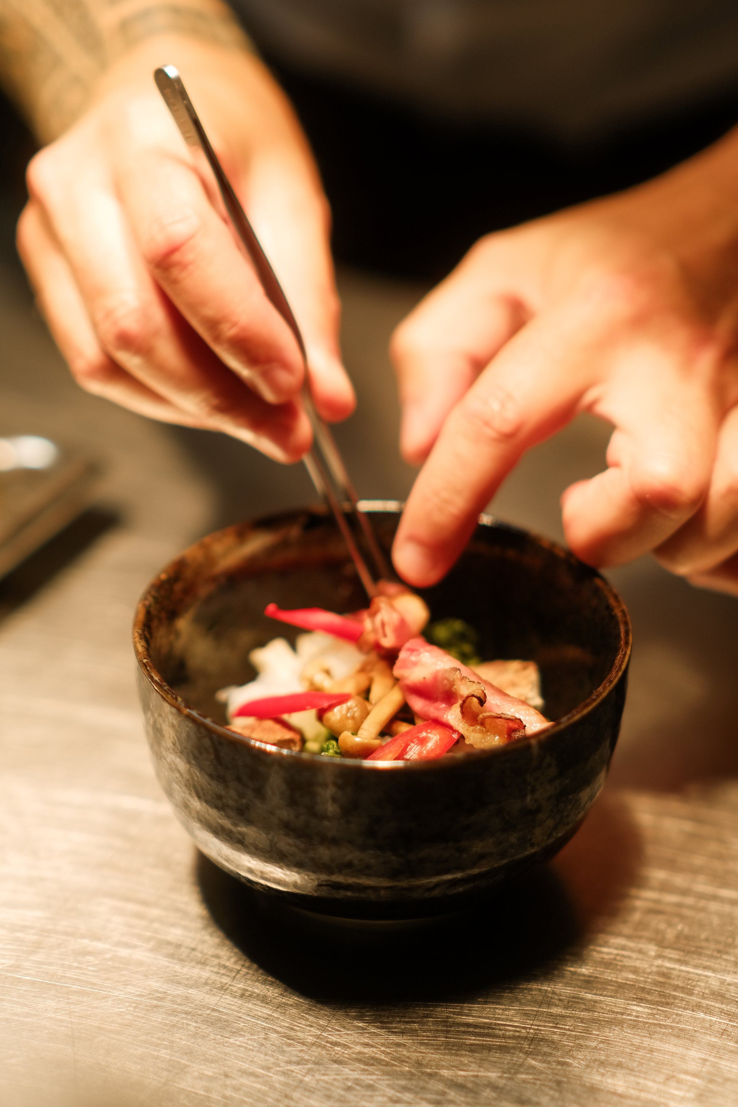 Nahaufnahme eines Kochs, der in einer schwarzen Schüssel mit Lebensmitteln wie Fleisch, Gemüse und Pilzen serviert und mit Zange und Finger arrangiert.