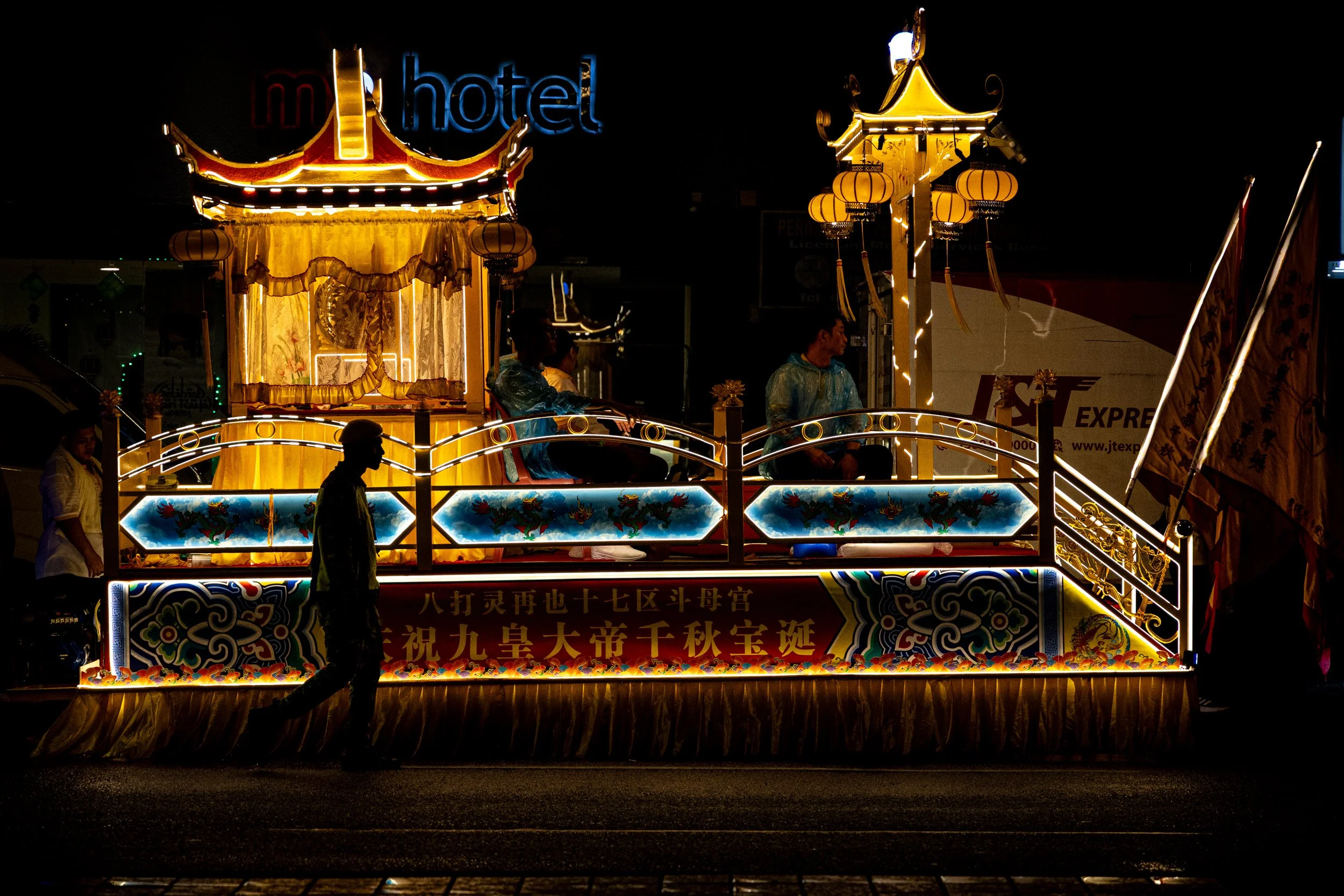 A religious procession float with Chinese characters glides through Jalan Pudu road in Kuala Lumpur at night with one man in the foreground and two men on the float.