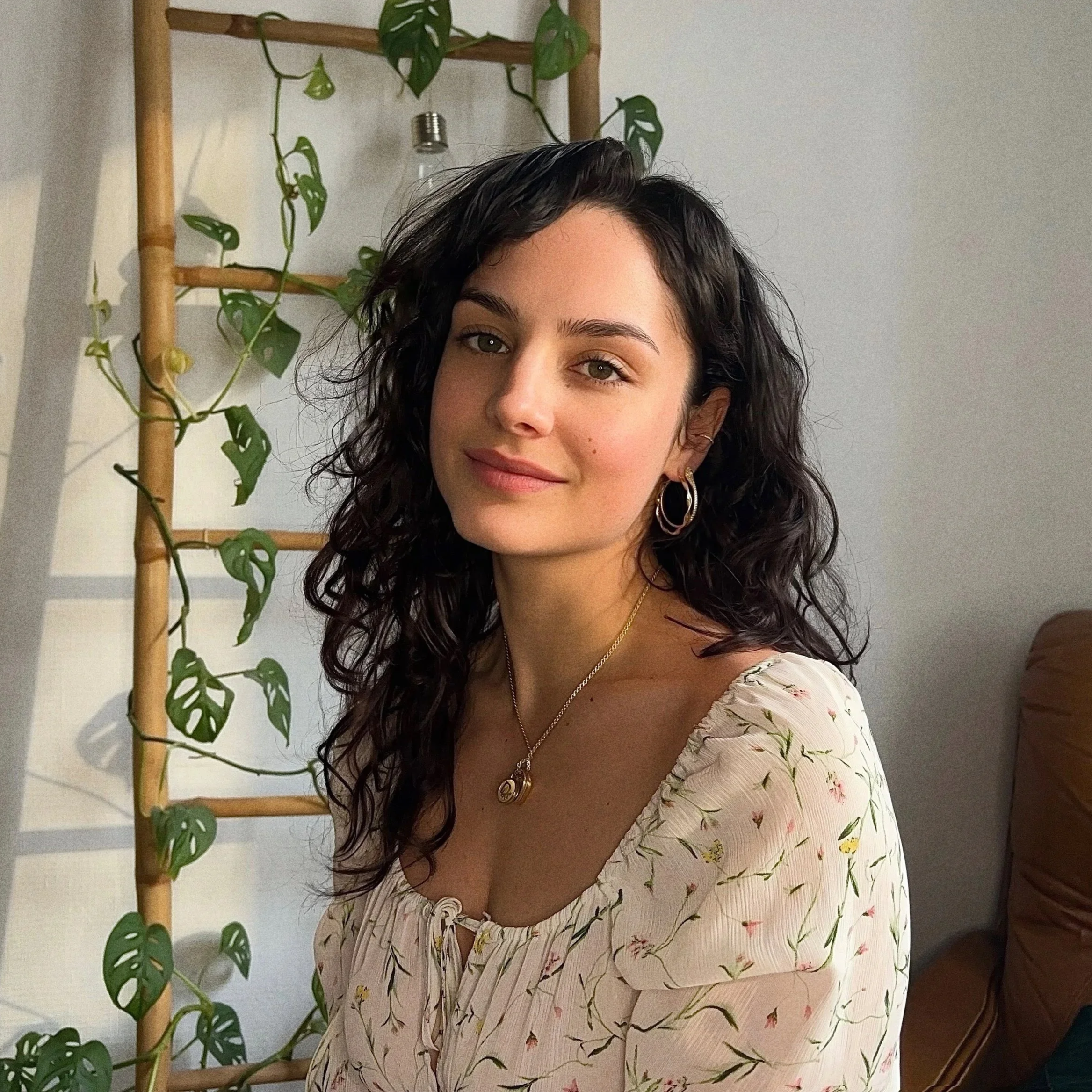 Une doula au sourire doux, avec des cheveux bouclés noirs porte une robe pâle à motifs floraux,  des bijoux en or, dans une pièce lumineuse avec une plante en arrière-plan.