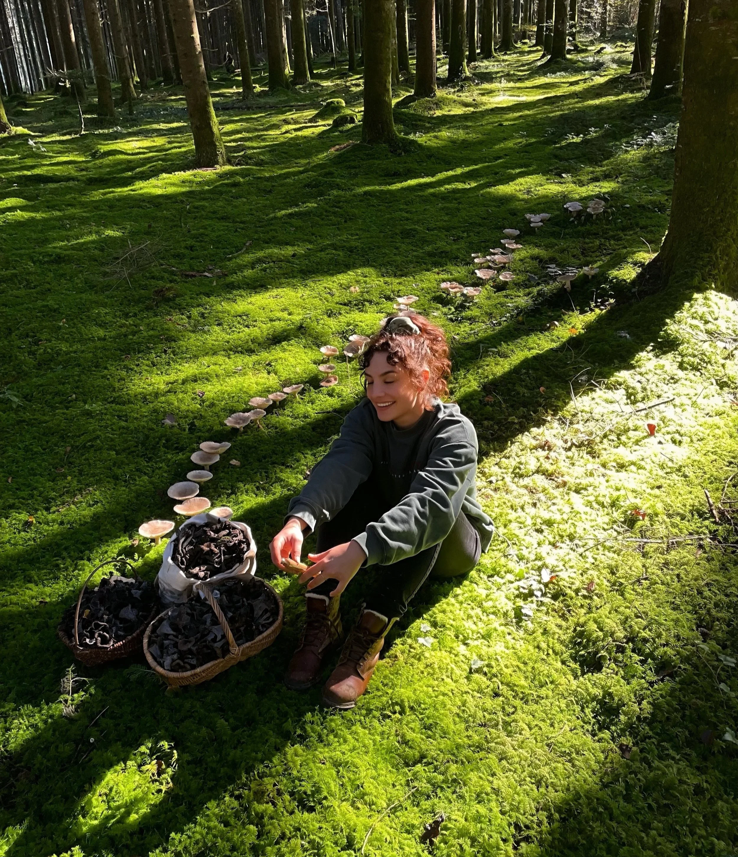 Pauline, une doula cueille des champignons dans une forêt verte ensoleillée de Mouchard dans le Jura, avec des paniers remplis de champignons autour d'elle.