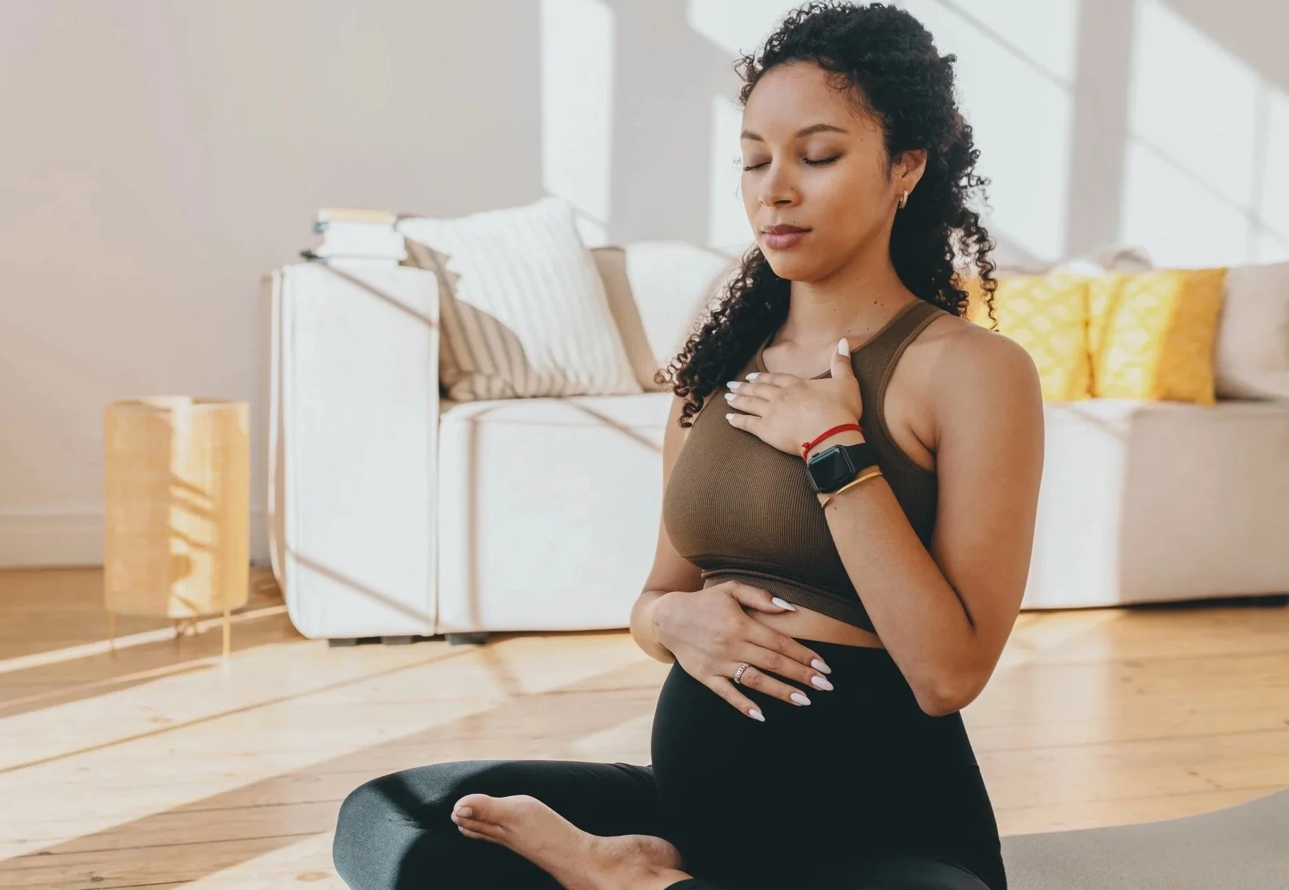 Une femme enceinte pratique des exercices de chant prénatal en position de lotus dans un salon lumineux du Jura, avec une main sur le cœur, portant une montre connectée et un bracelet, vêtue d'un haut marron et de leggings noirs.