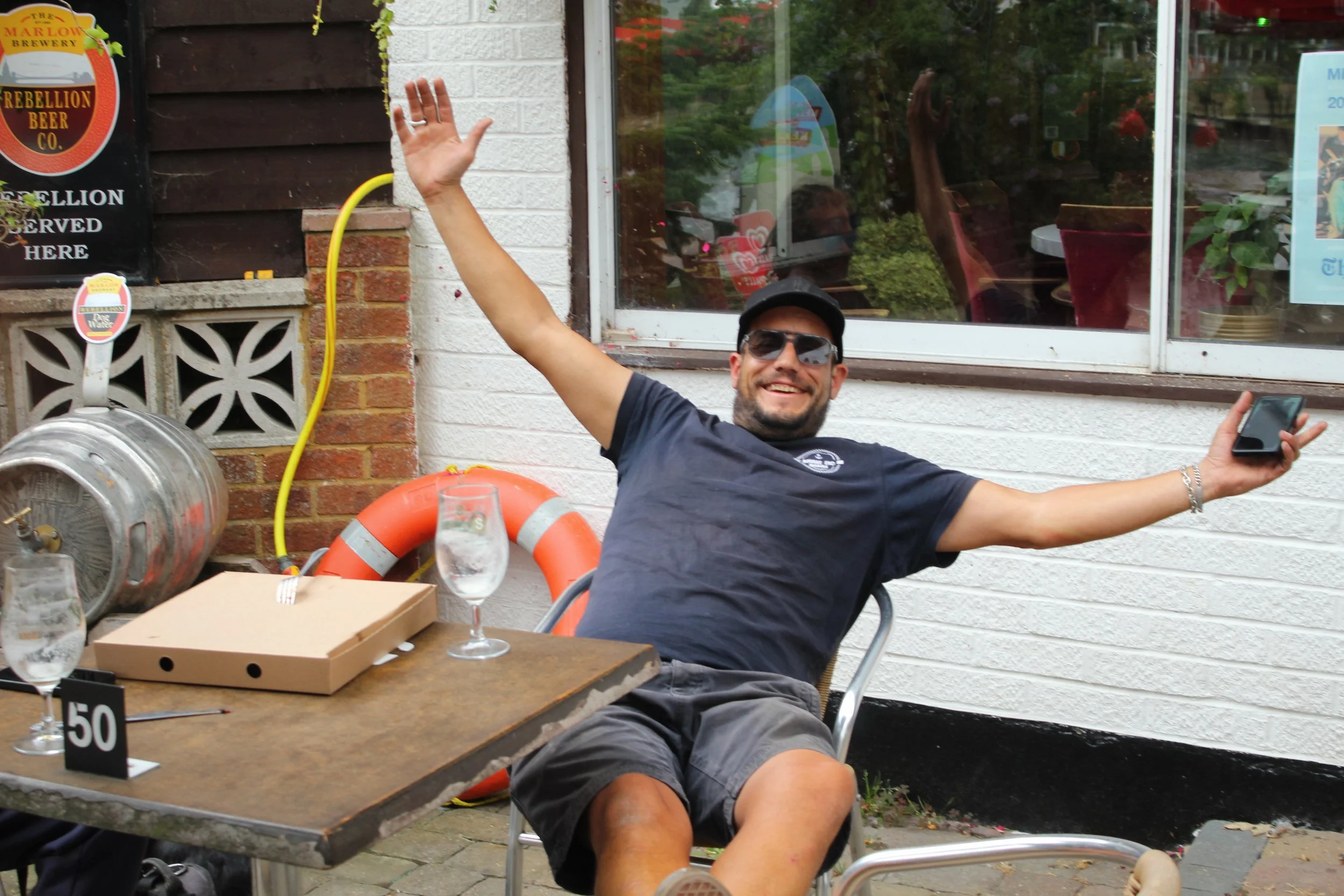 A man wearing sunglasses, a black cap, and a dark t-shirt sitting outdoors at a table. He is smiling broadly with his arms raised in a celebratory gesture. On the table are two glasses of water, a pizza box, and a black number sign with the number 50. The background shows a building with a white brick and dark wood exterior, a yellow hose, an orange life preserver, and a window reflecting trees and sky.