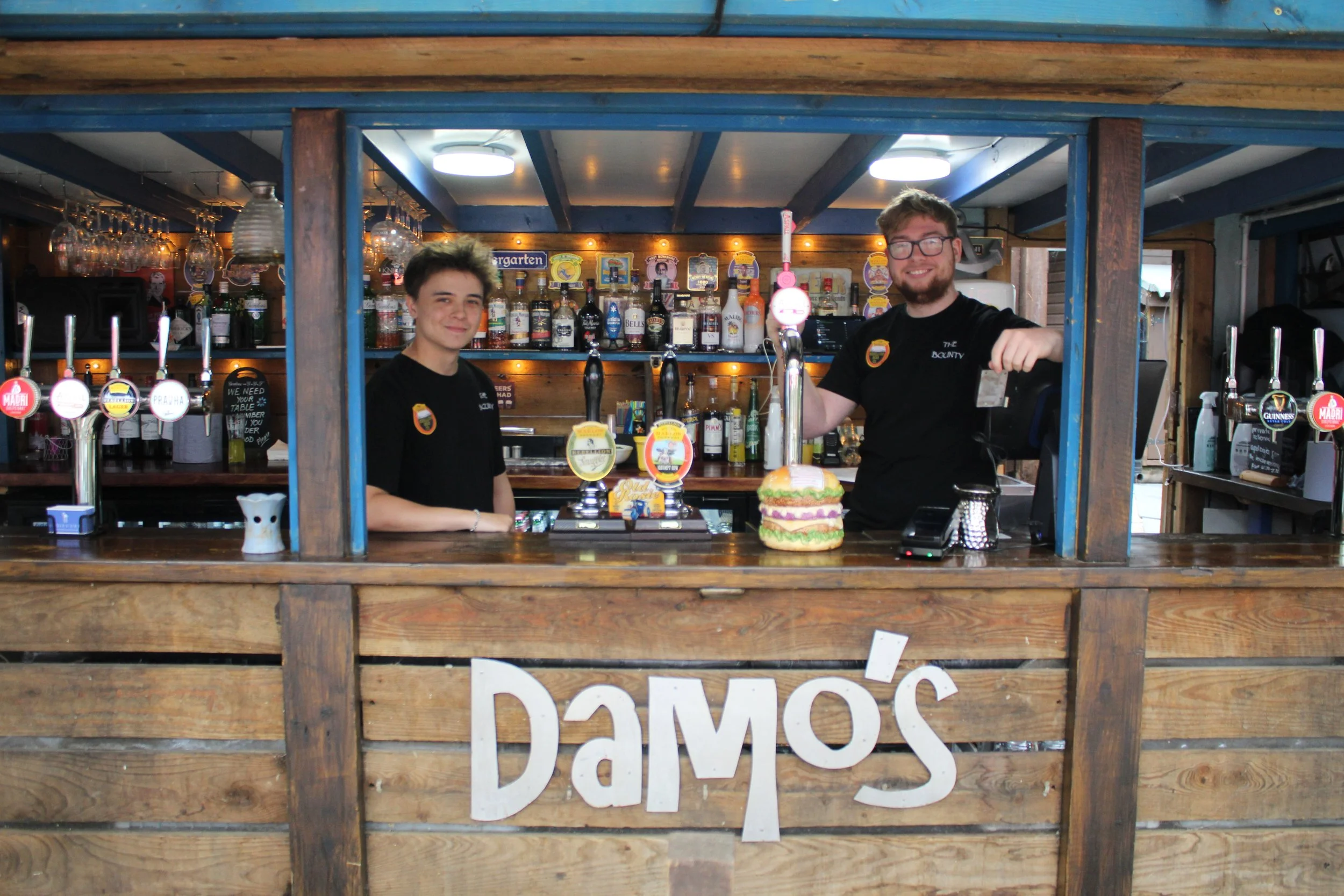 Two young men working behind a rustic wooden bar with a sign that reads 'Damos'. The bar has multiple beer taps, bottles of alcohol, and a large burger prop on the counter. The background includes shelves with liquor bottles and glassware, indicating