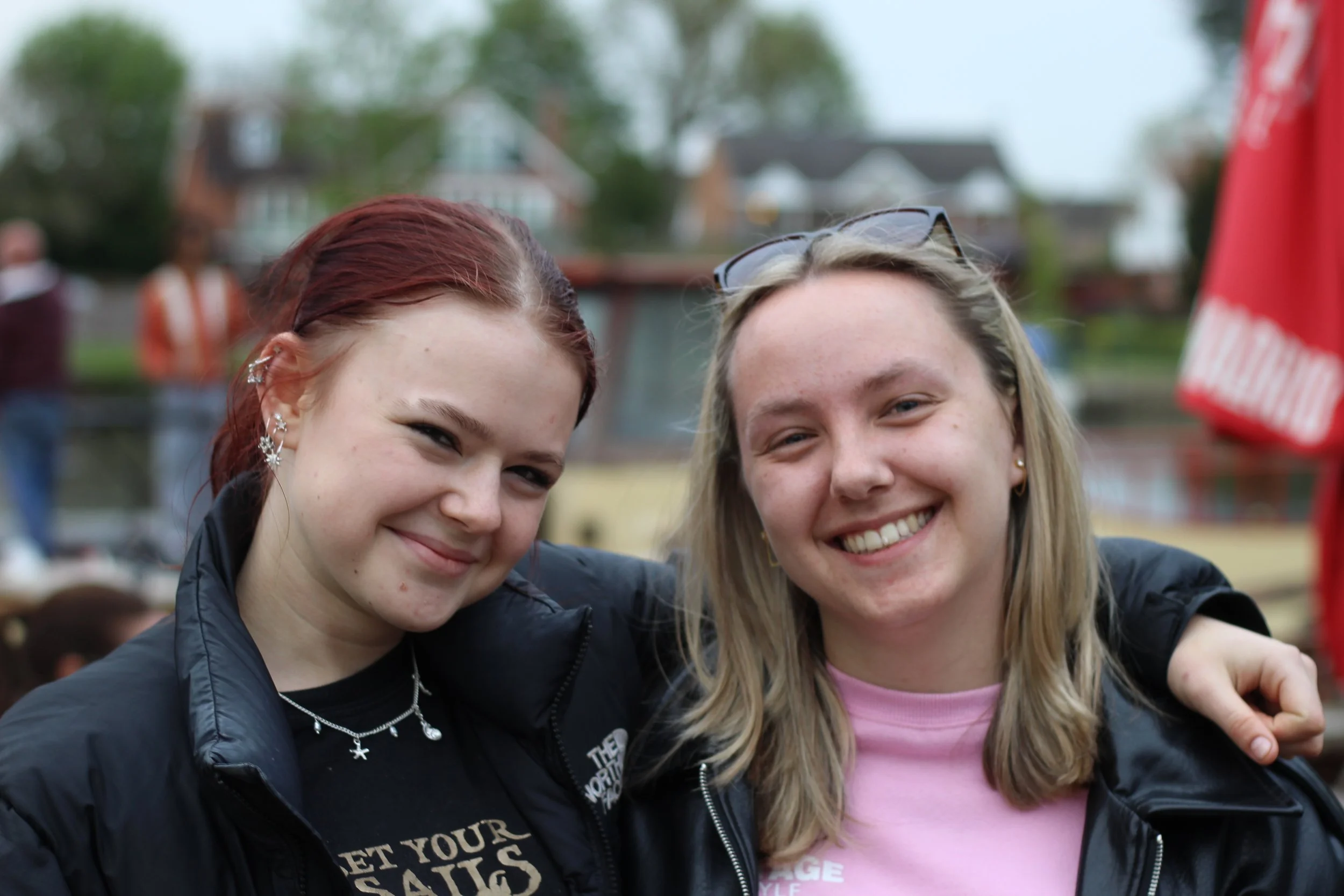 Two young women smiling with their arms around each other, outdoors on a cloudy day, with blurred trees and houses in the background.