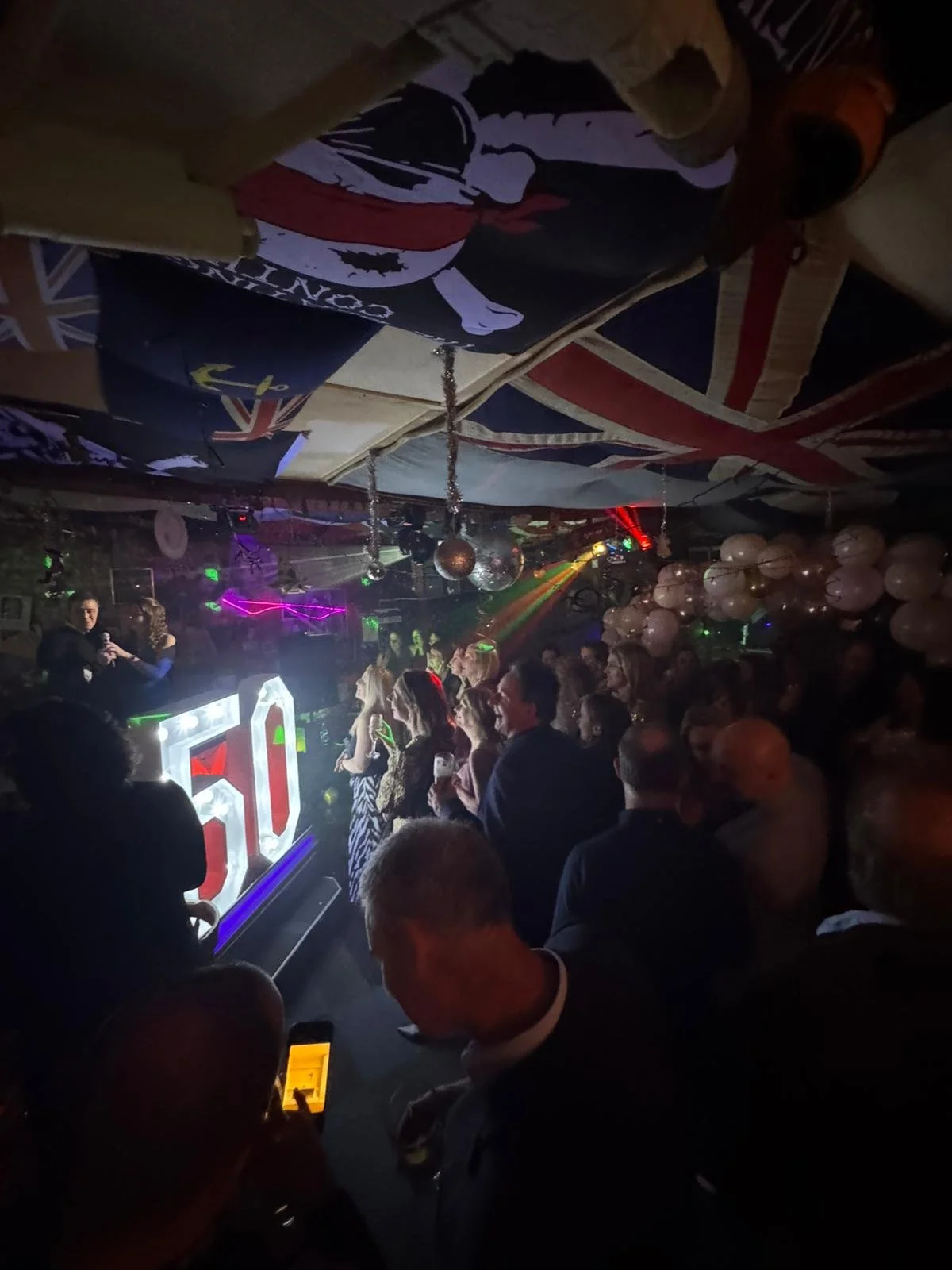 A lively nightclub scene with people dancing and socializing near a DJ booth with large illuminated '50' sign, decorated with Union Jack flags and balloons, with disco balls and colorful stage lights overhead.