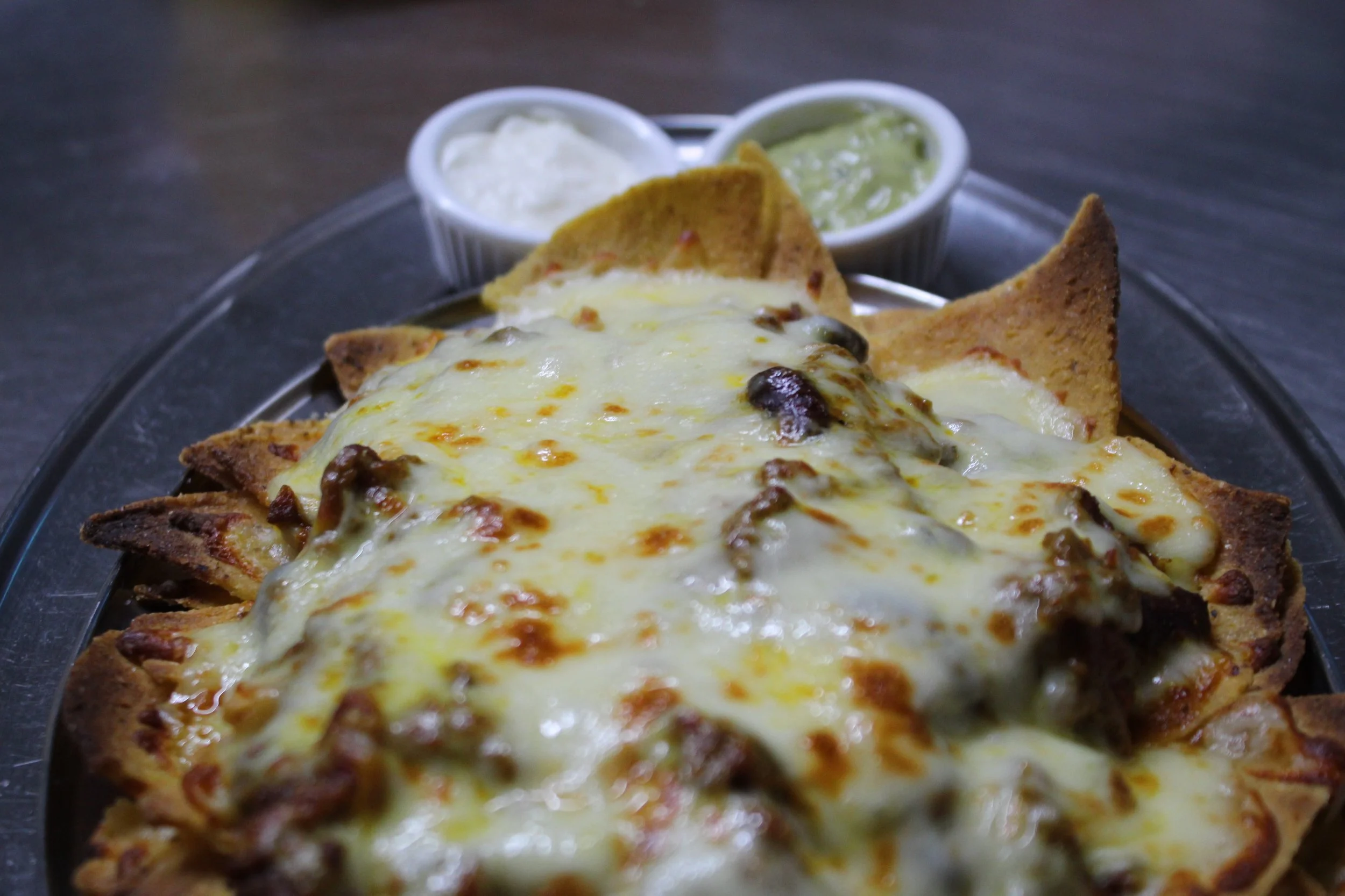 Cheeseburger nachos topped with melted cheese and jalapeños, served with two small bowls of sour cream and guacamole in the background.