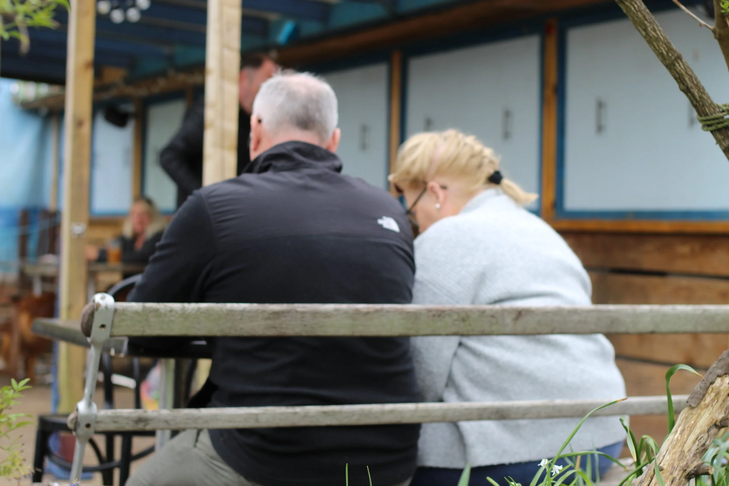 A man and woman sitting on a bench outdoors, leaning in close, with several people visible in the background at what appears to be a casual dining or social setting.