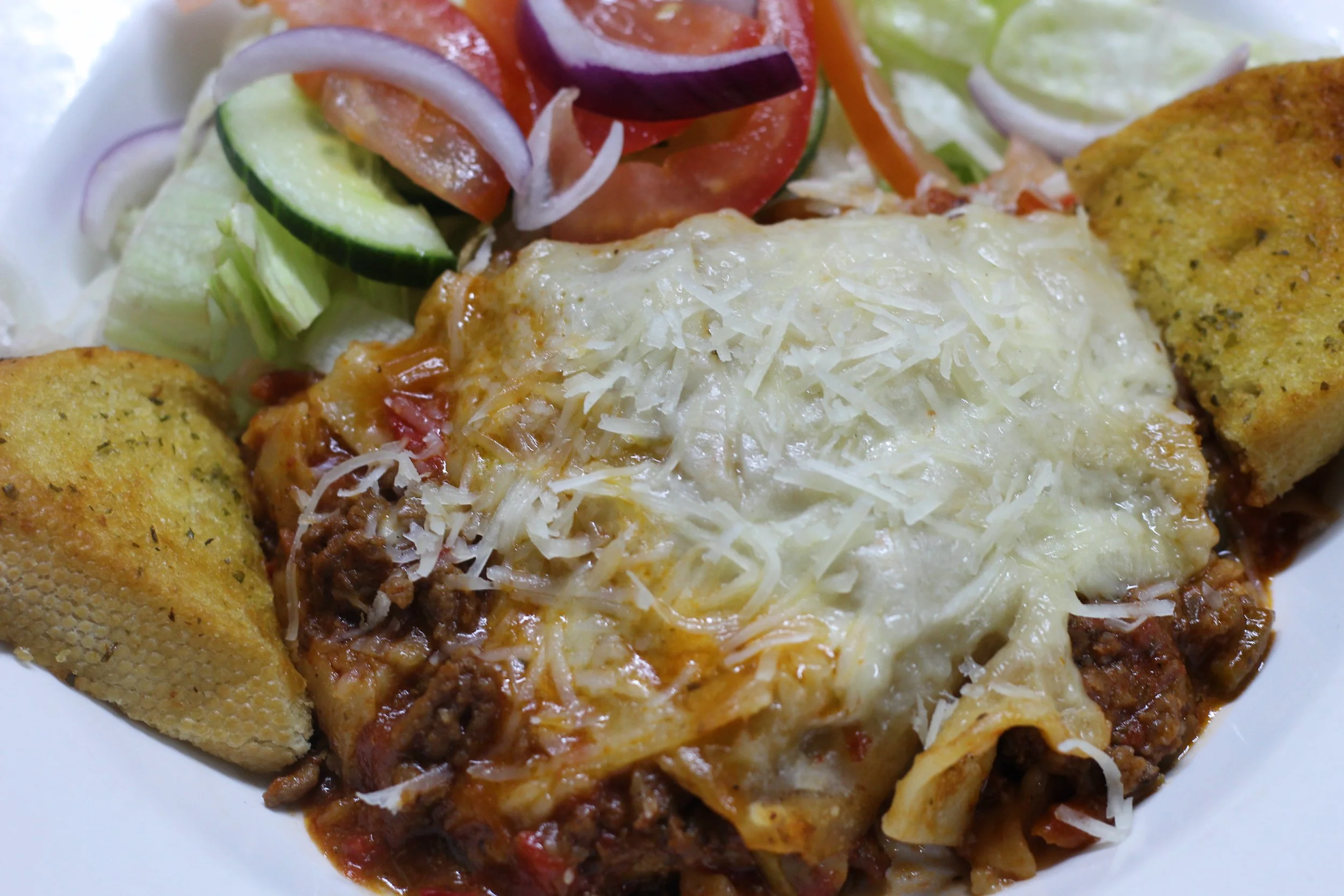 A plate of baked lasagna with melted cheese, served with side of salad containing sliced cucumbers, tomatoes, onions, and shredded lettuce, with two pieces of garlic bread.