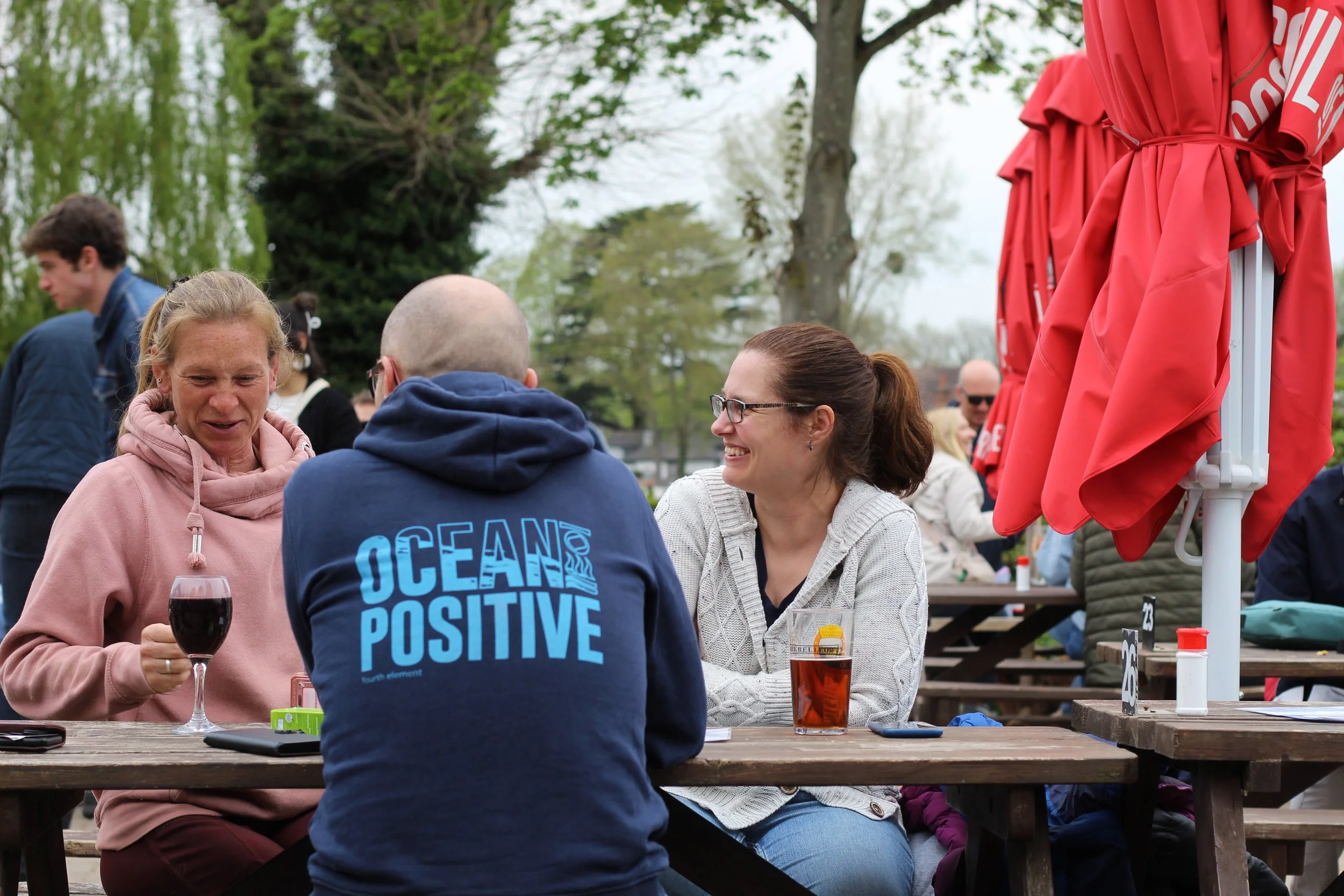 Three people sitting at a wooden outdoor table, talking and smiling, with drinks in front of them, during daytime at a park or outdoor event.