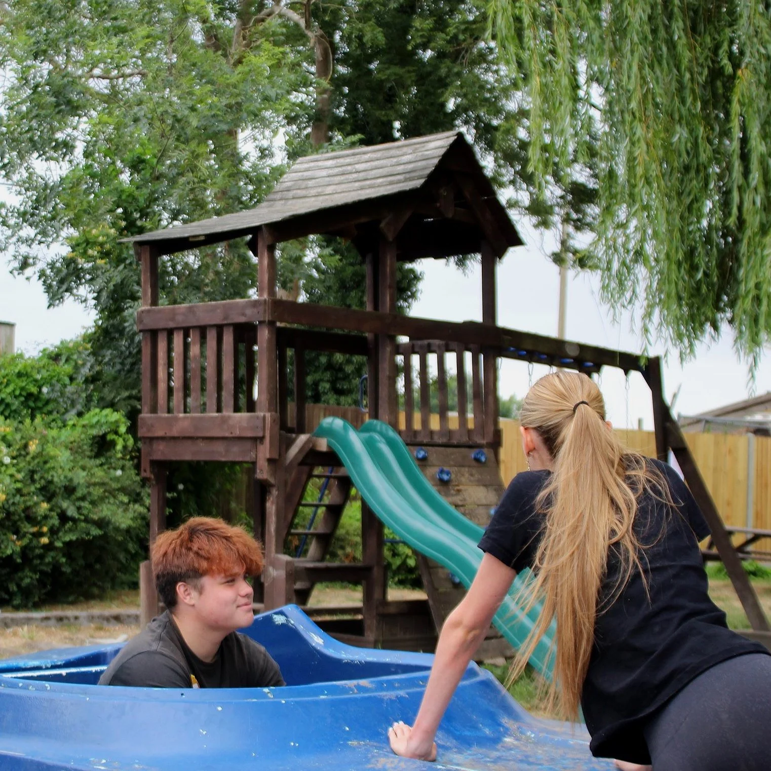 Two teenagers near a plastic kiddie pool with a wooden play structure and green slide in the background, outdoors among trees and plants.