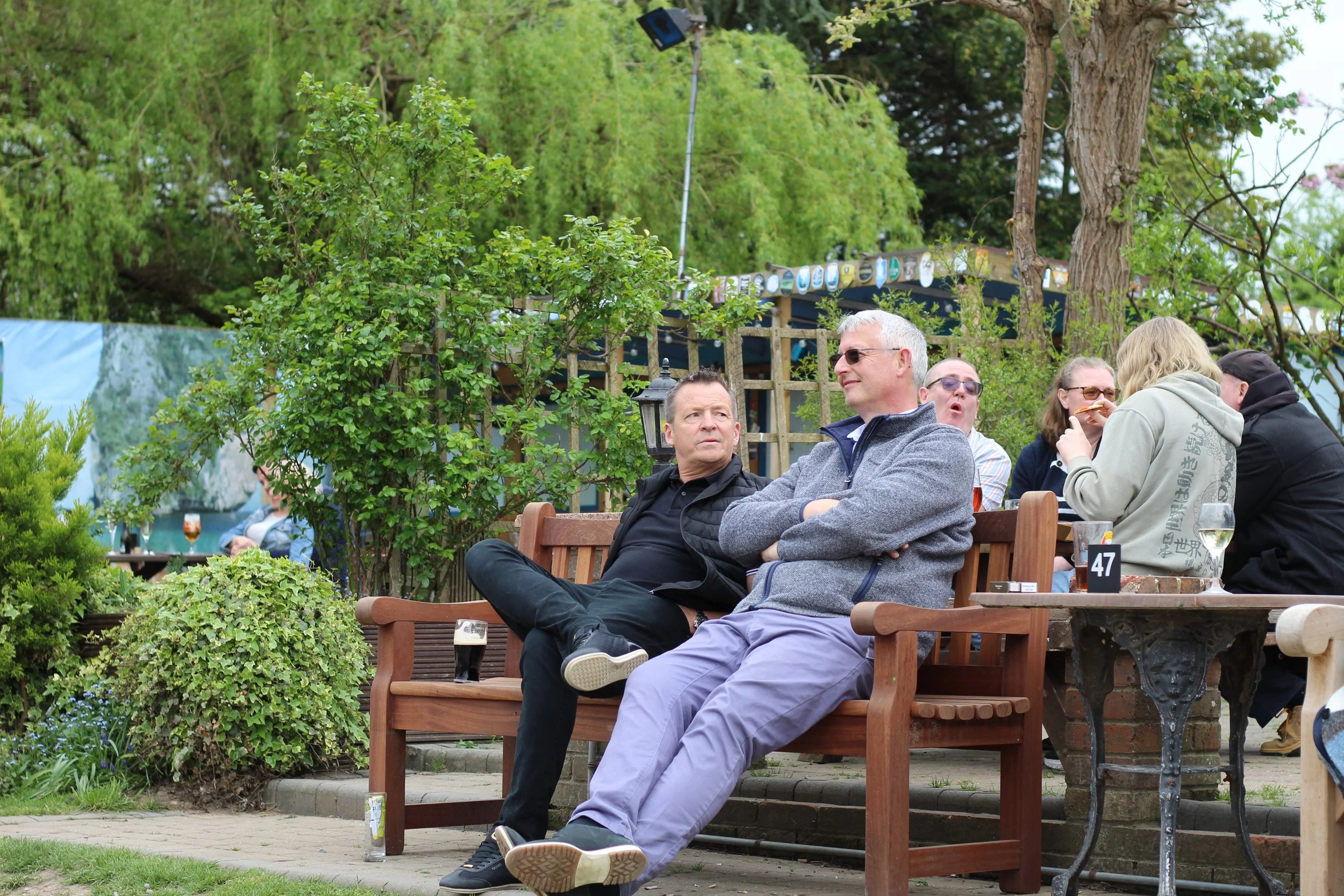 People sitting on a wooden bench and outdoor table at a garden party, with drinks, surrounded by greenery.