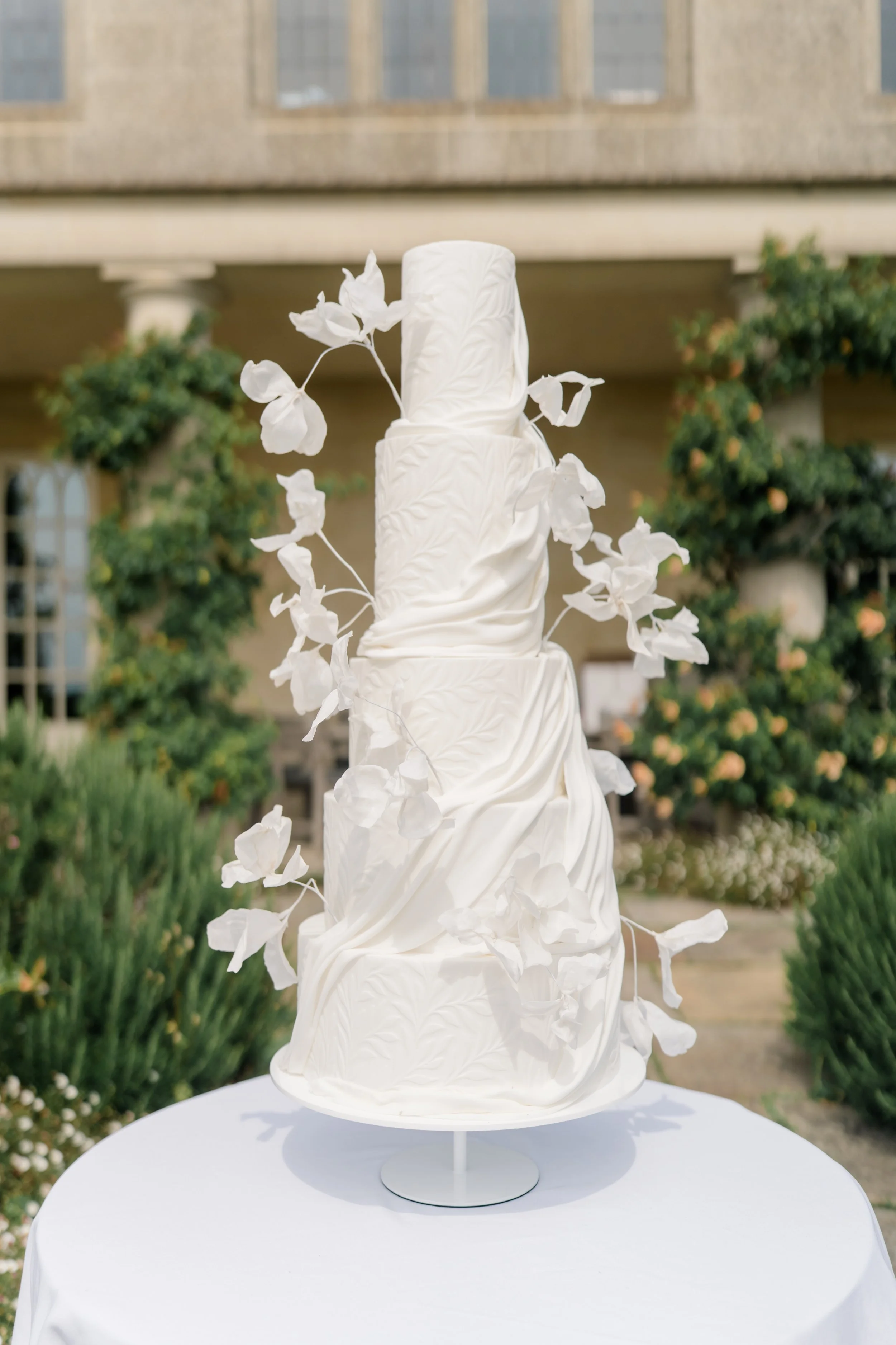 White wedding cake with a textured, draped design, decorated with white sugar flowers and leaves, displayed outdoors on a round white table. Styled shoot held at Euridge Manor assisted by Bootique Weddings and Events.