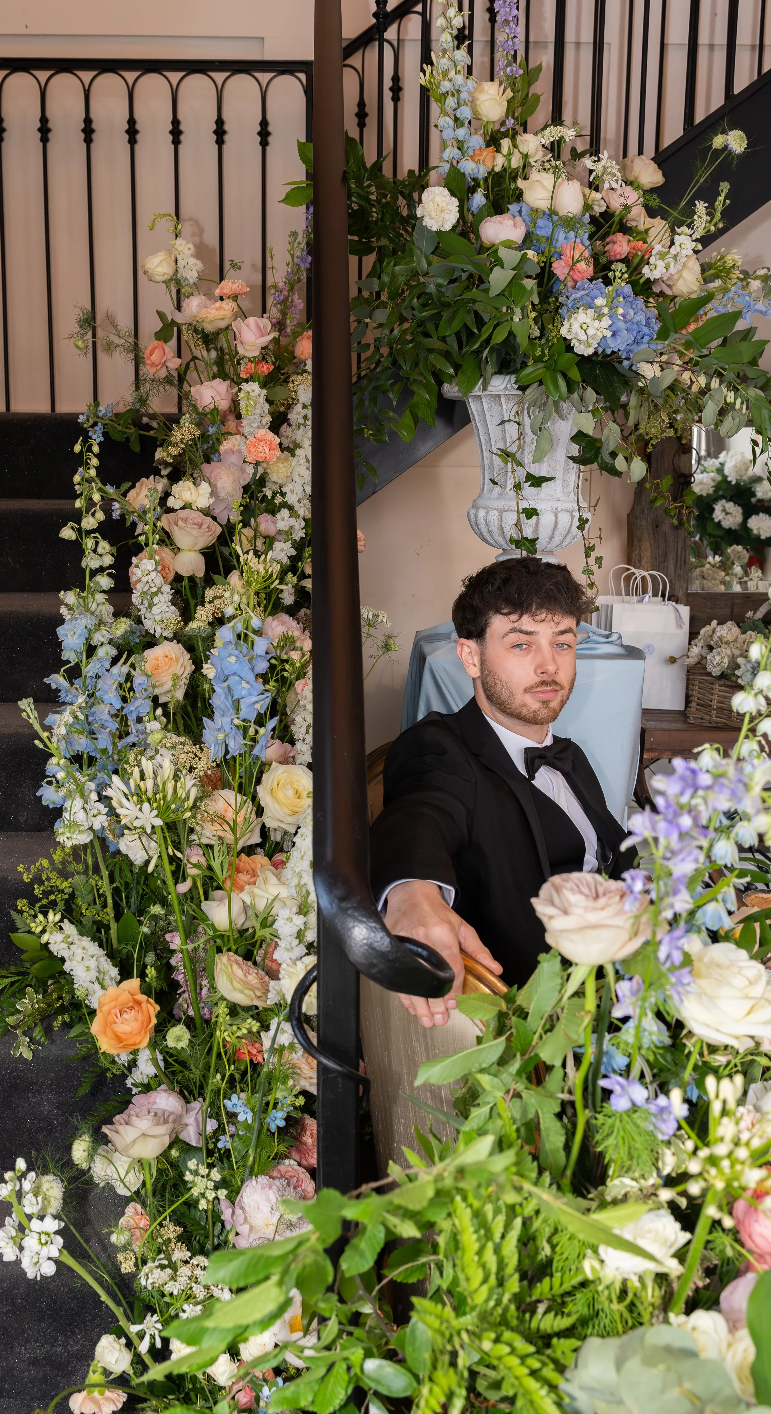A man in a tuxedo sitting on a chair surrounded by an arrangement of colorful flowers, with floral decorations on a staircase and a large floral arrangement in a white vase nearby. Styled shoot held at The Cheshire Venue.