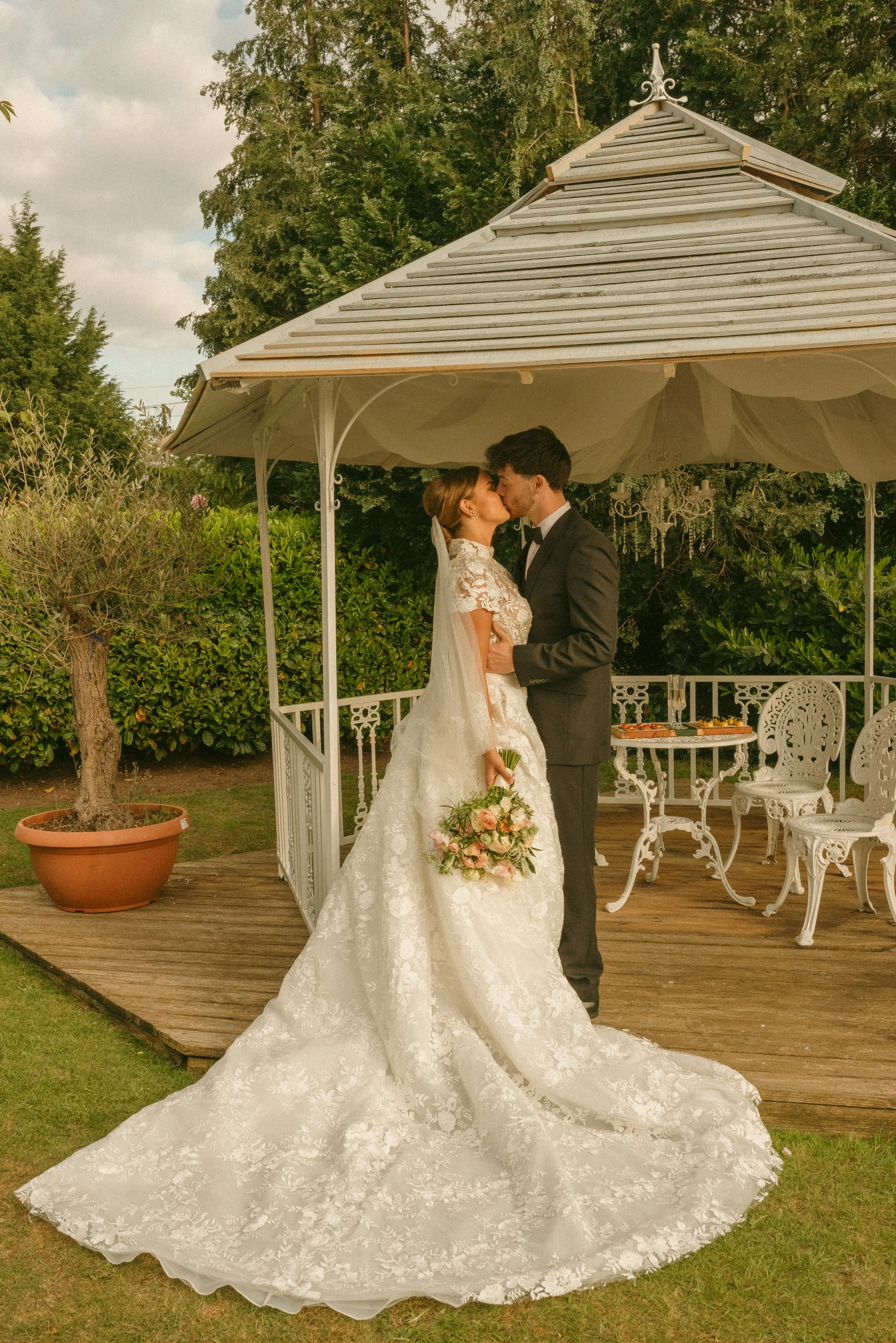 Bride and groom sharing a kiss outdoors under a gazebo, surrounded by greenery, with wedding attire and a bouquet of flowers. Styled shoot held at The Cheshire Venue planned by wedding planner Bootique Weddings and Events.