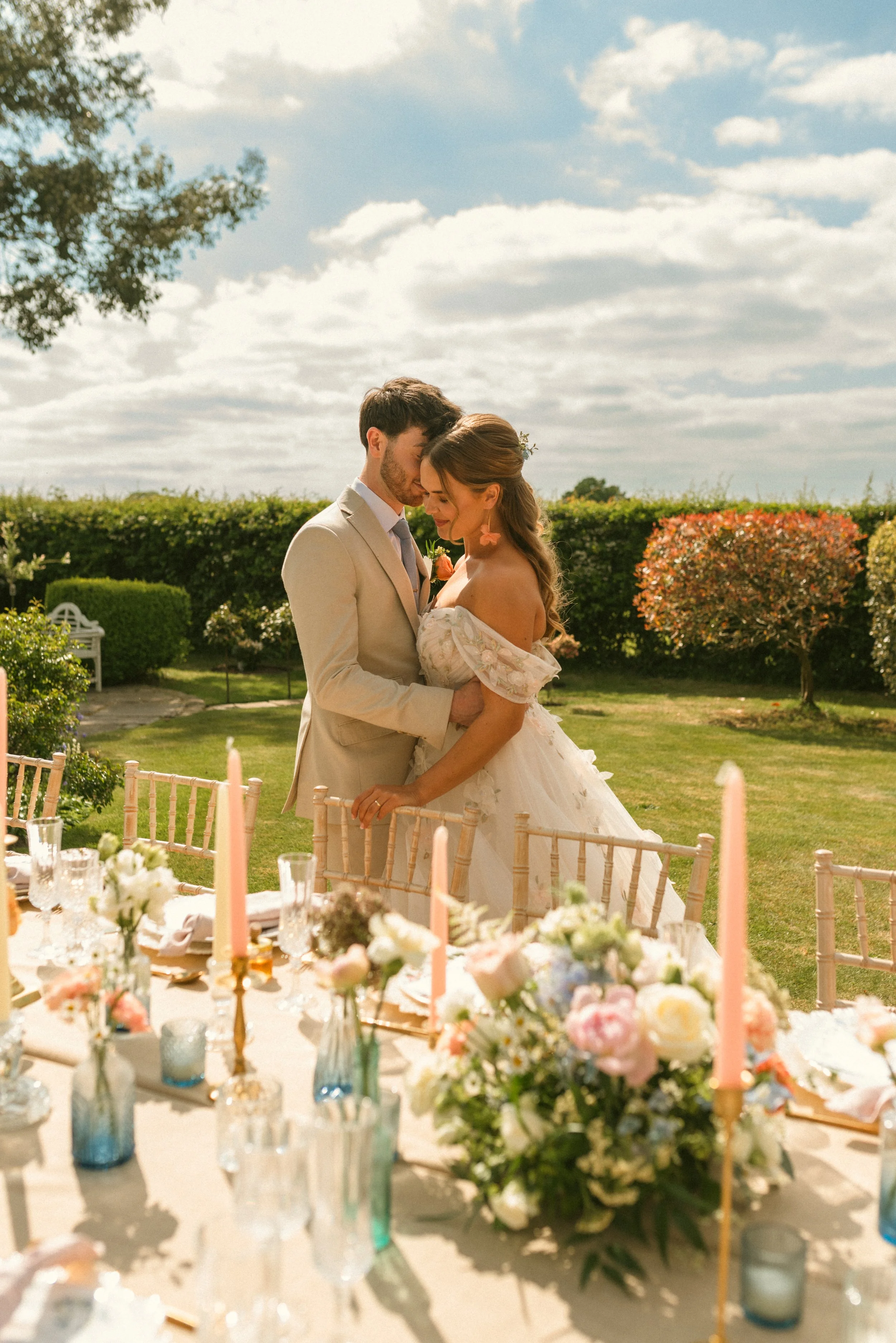 A bride and groom embrace in a garden during their wedding, surrounded by floral arrangements and set against a bright, partly cloudy sky. Taken at a styled shoot at The Cheshire Venue and planned by Bootique Weddings and Events.